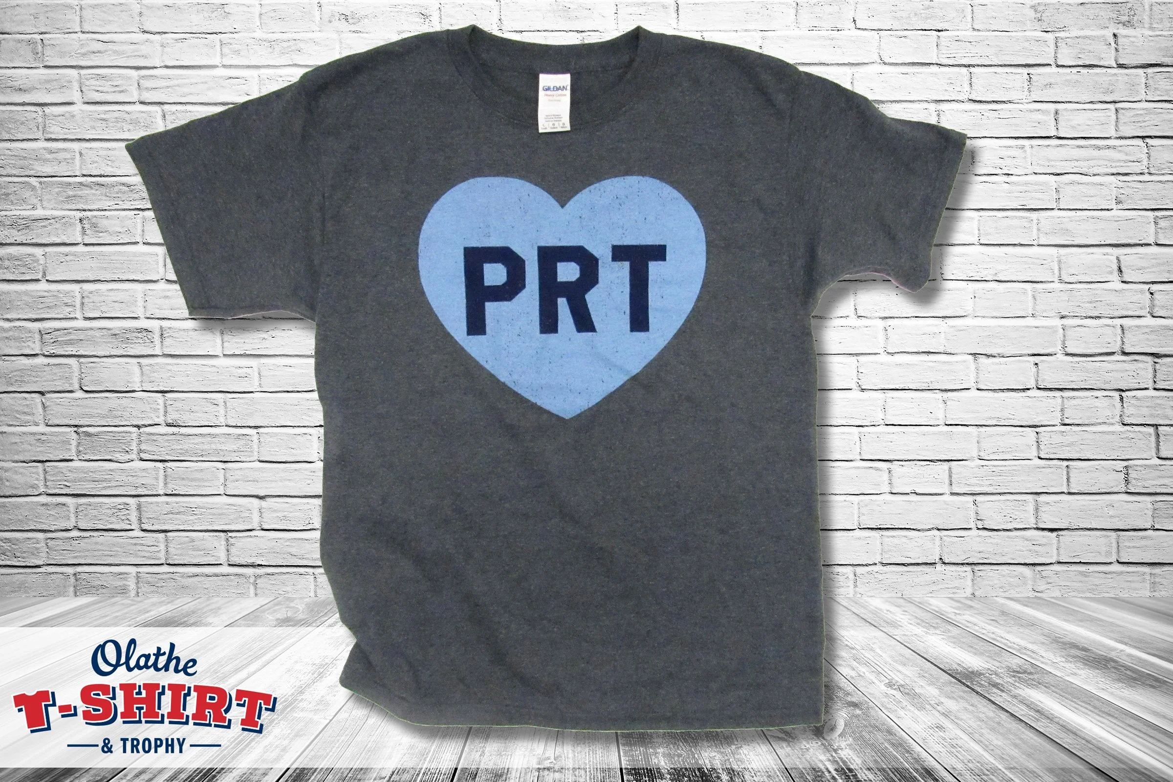 Gray T-shirt with a large blue heart design and the letters "PRT" inside the heart, hanging on a white brick wall.