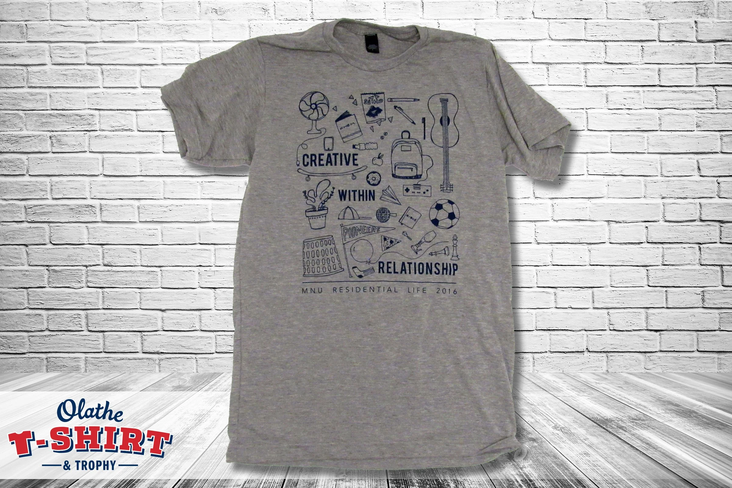 Gray T-shirt with illustrations and decorative lettering, displayed against a white brick wall.
