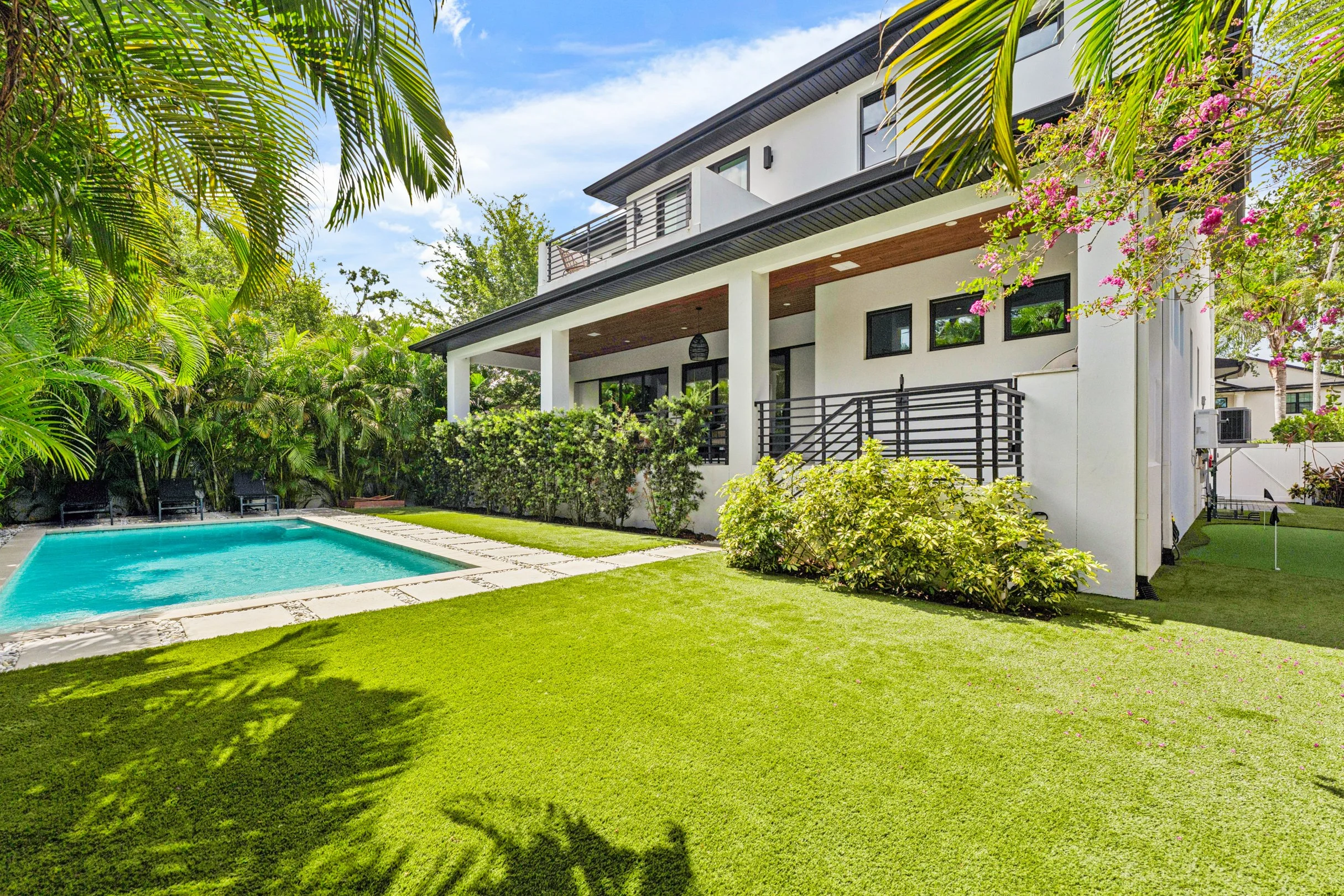 Modern two-story house with a white exterior, black window frames, front porch, surrounded by lush green lawn, tropical plants, and trees, with a rectangular swimming pool and outdoor lounge chairs.