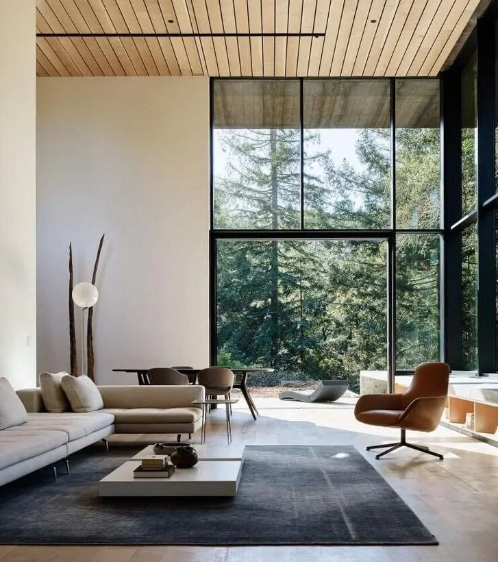 Modern living room with a large floor-to-ceiling glass window showing trees outside, light wood ceiling, beige sectional sofa, orange armchair, black dining table with chairs, and a white coffee table on a dark rug.
