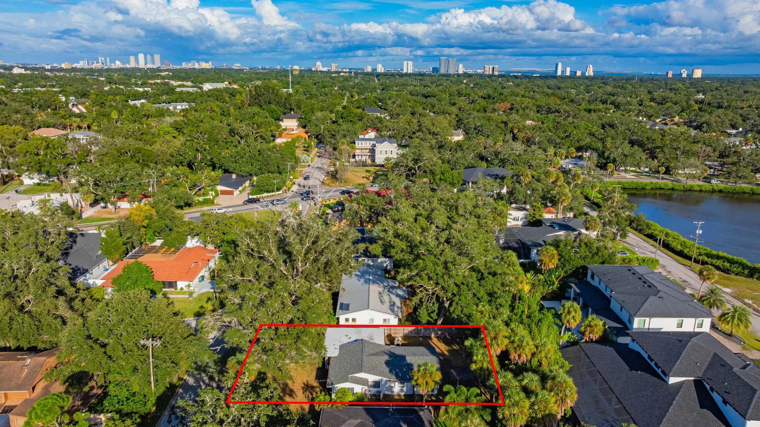 Aerial view of a suburban neighborhood with a single-family house outlined in red, surrounded by trees and adjacent to a small pond, with a city skyline in the distance under a partly cloudy sky.