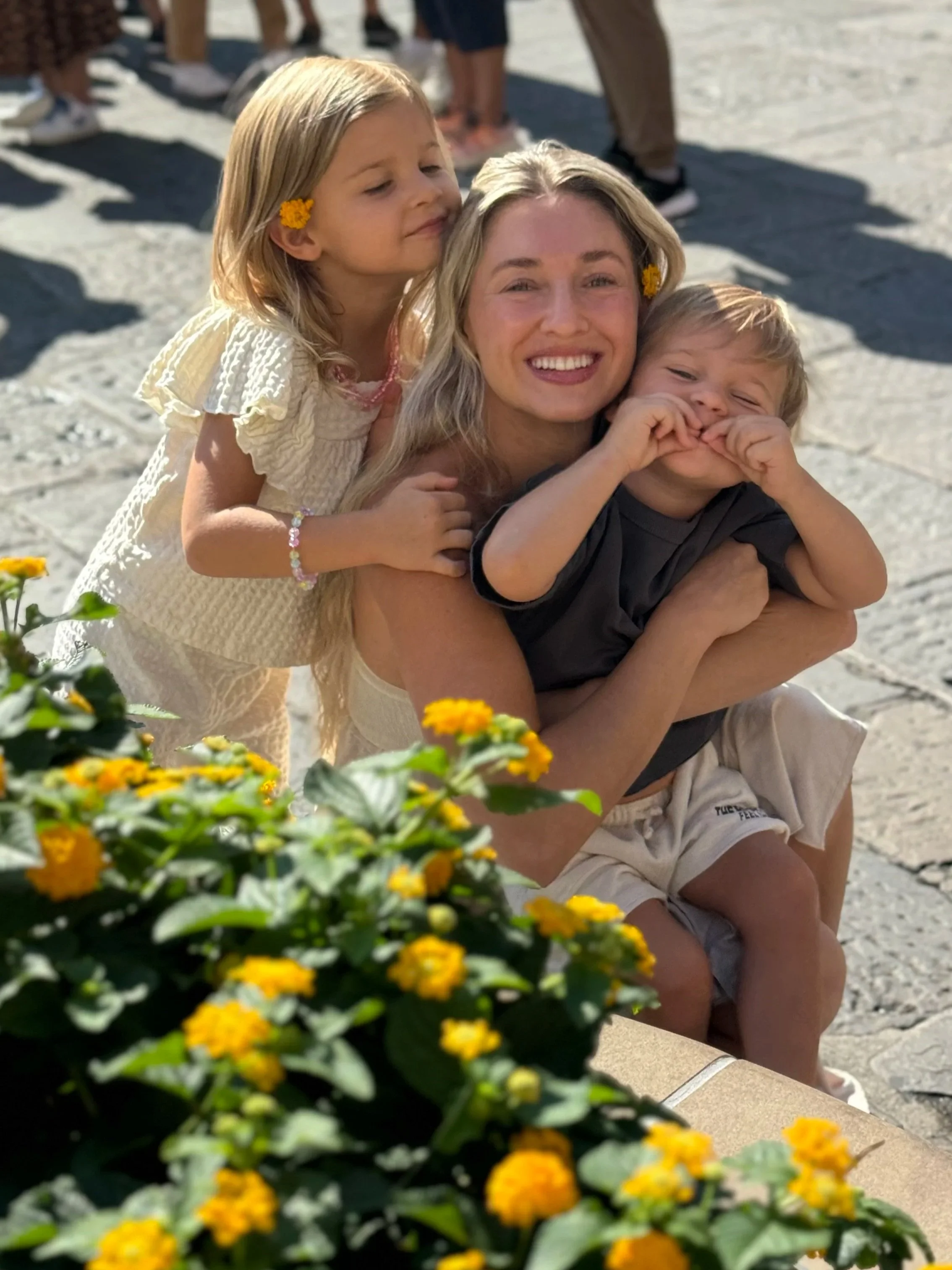 A woman and two children smiling on a sunny day, with yellow flowers in the foreground.
