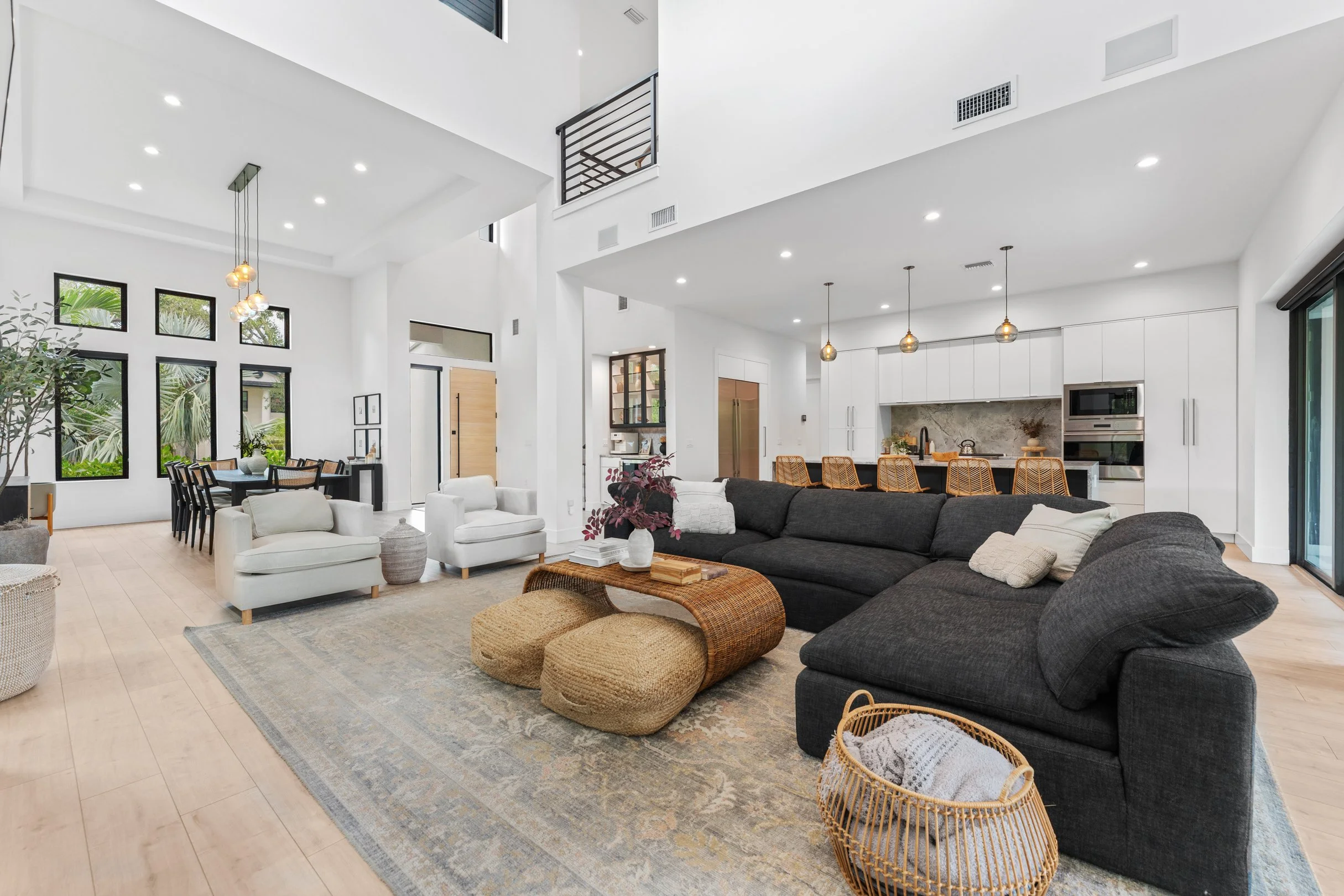 Open-concept living room with white walls, high ceilings, and large windows, featuring a black sectional sofa, two white armchairs, a woven coffee table, and a dining area in the background.