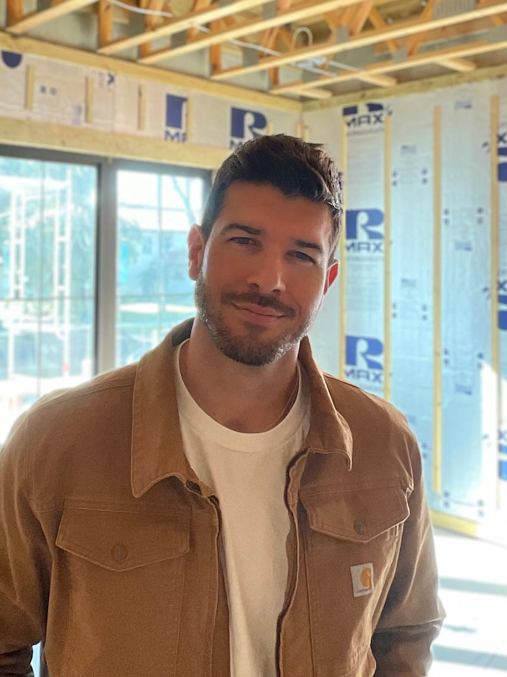A man with dark hair, beard, and mustache smiling at the camera in a construction site with insulation and wooden beams.