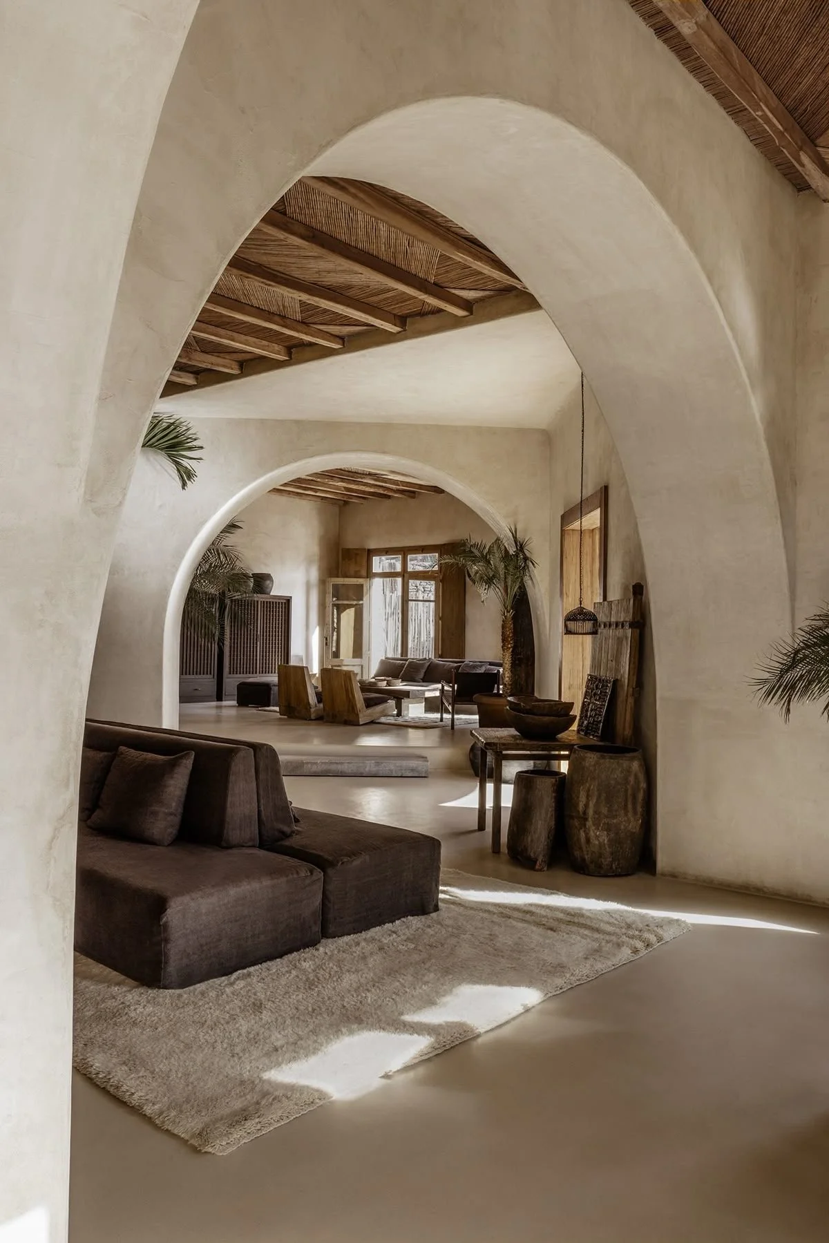 A spacious living room with beige textured walls, arched doorways, and wooden ceiling beams, furnished with contemporary sofas, chairs, and decorative wooden tables, accented by potted plants and natural sunlight.