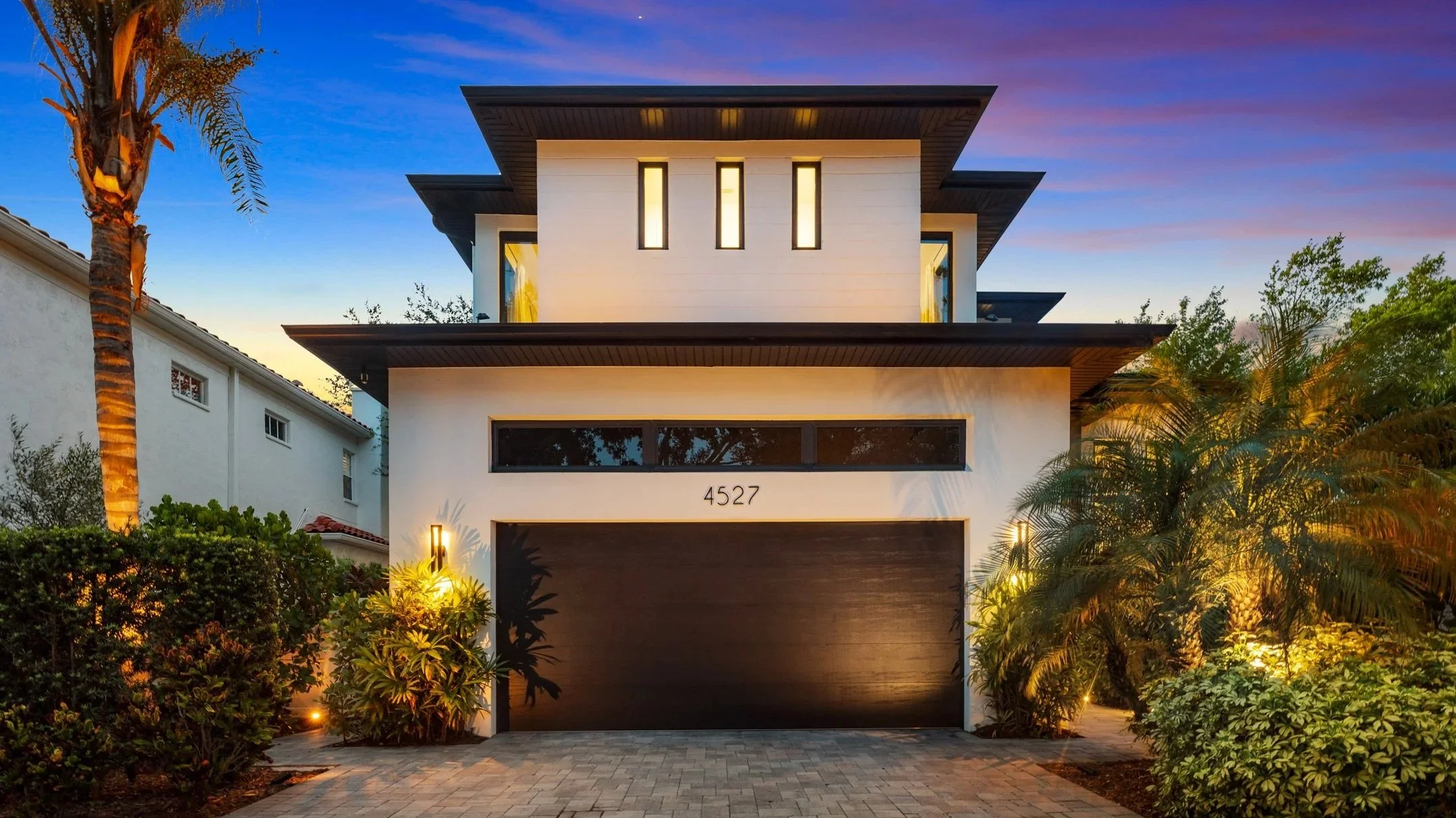 Modern two-story house with a black garage door, illuminated exterior lights, tropical landscaping, and large windows, under a colorful evening sky.