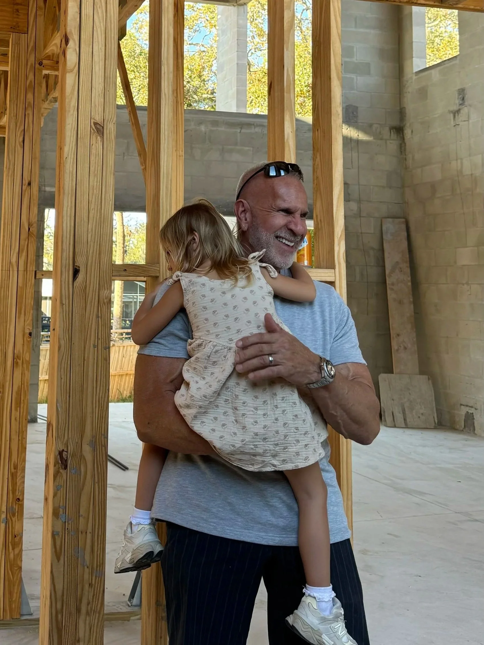 A smiling man holding a young girl in a construction site with wooden framing around them.