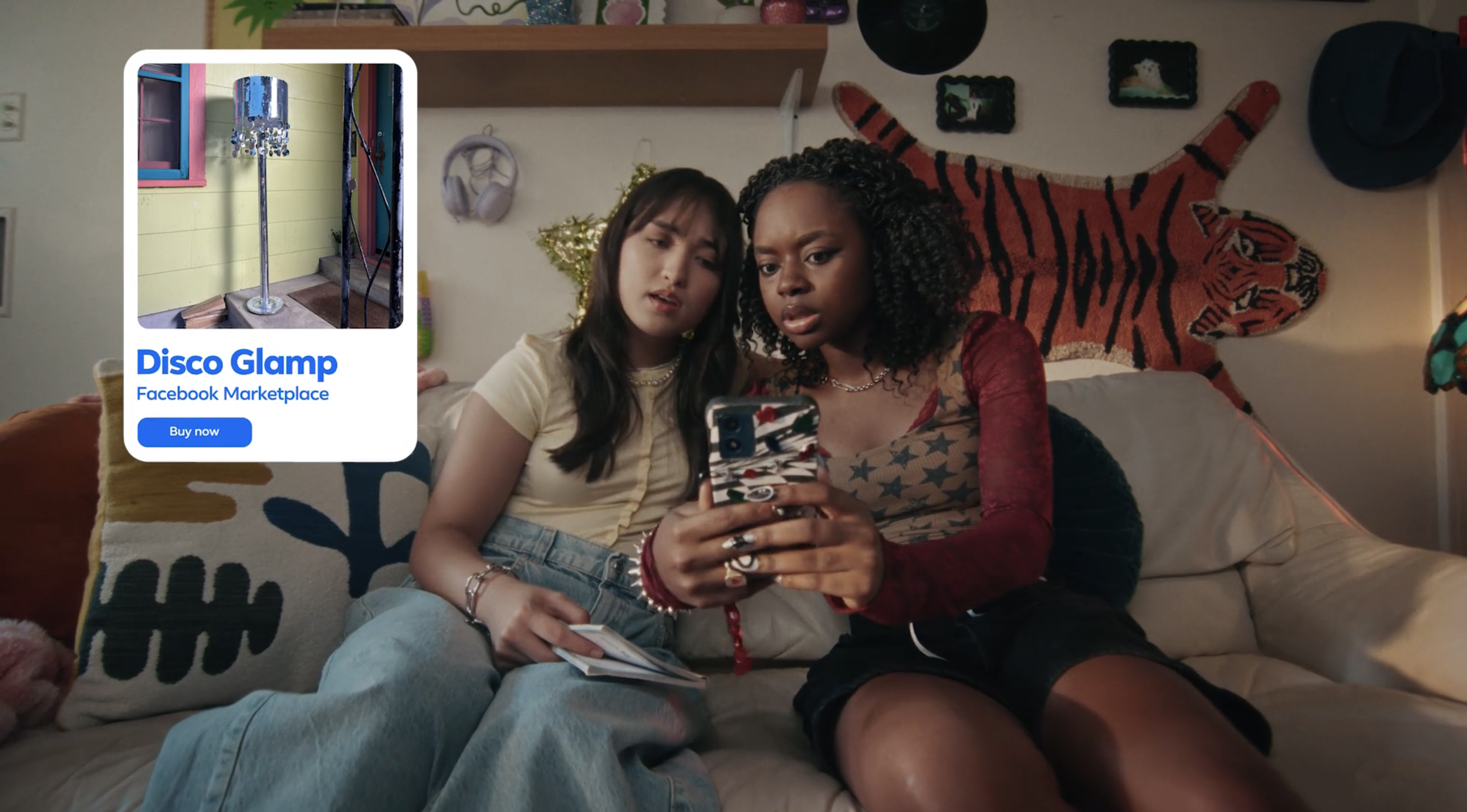 Two young women sitting on a beige couch, looking confused at a smartphone with a tiger print case. Behind them is a wall decorated with a tiger tapestry and various pictures. The woman on the left has long dark hair, wears a yellow top, and has a no