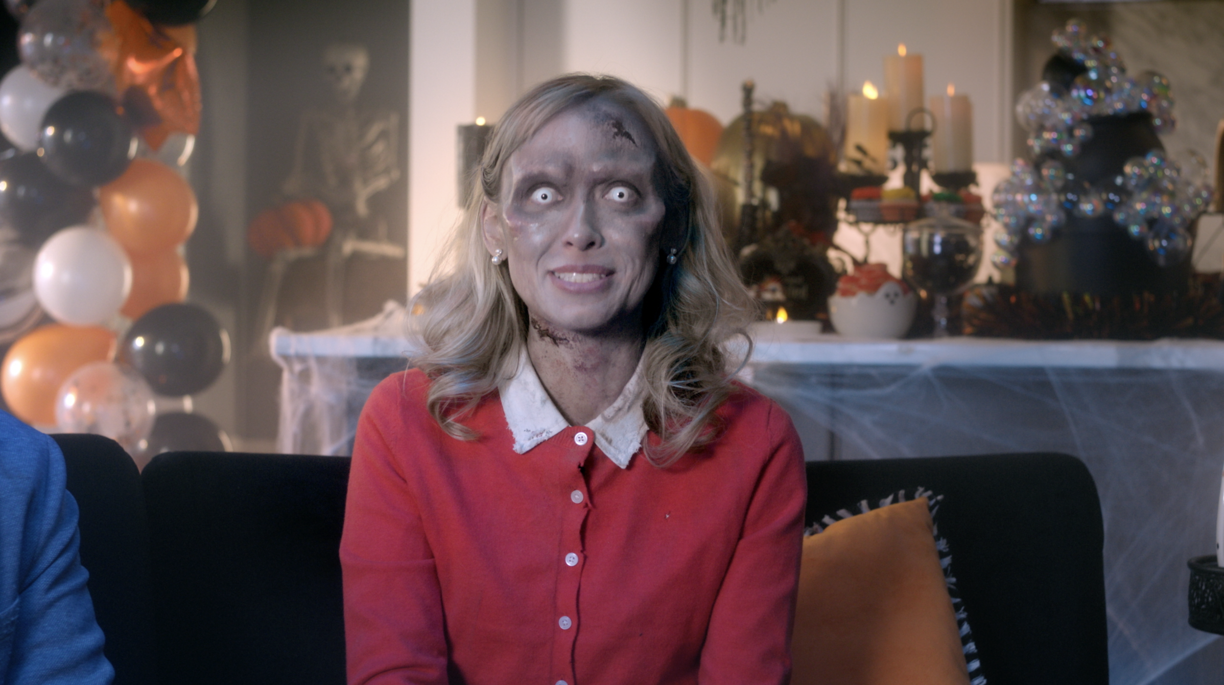 A woman with zombie makeup and pale, glowing eyes sitting on a couch at a Halloween party, decorated with balloons, cobwebs, and Halloween-themed decorations.