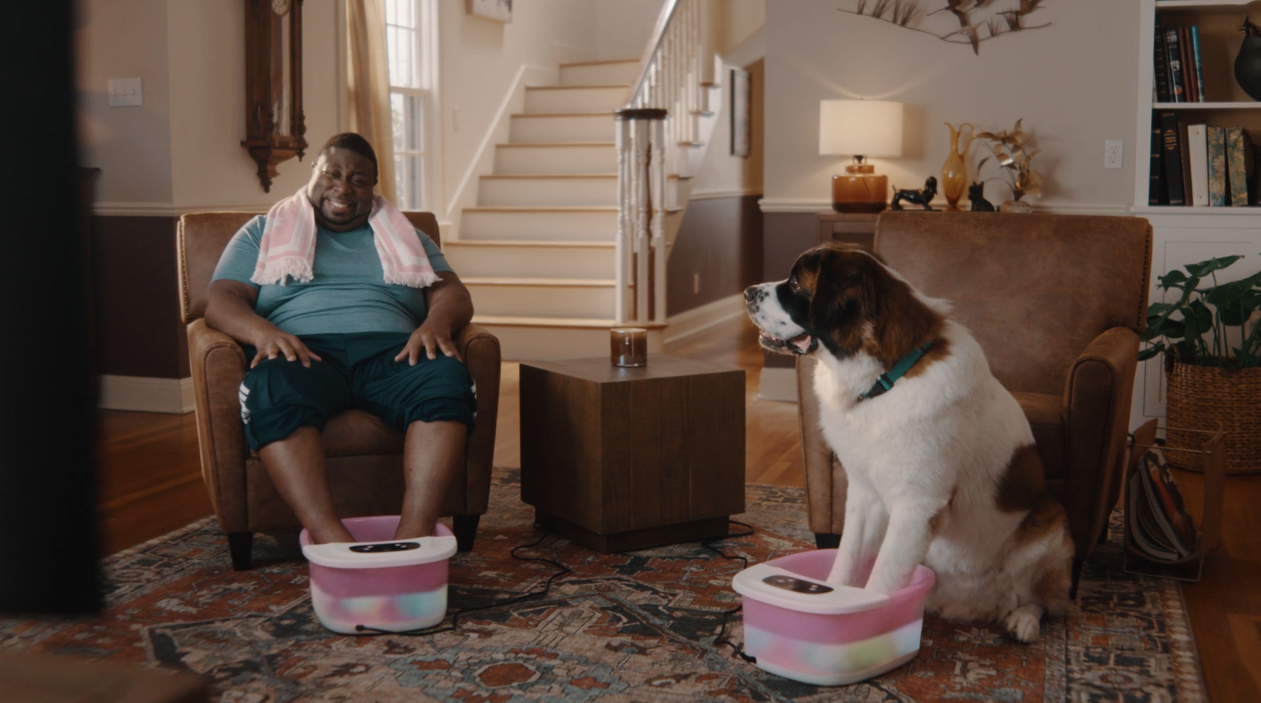 A man with a towel around his neck is sitting in a chair with his feet in a pink foot soak basin, smiling at a dog with a similar basin, as if having a spa day at home.