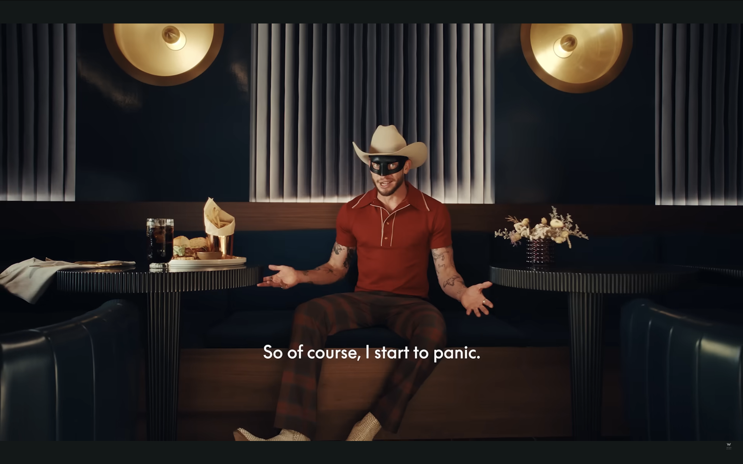 A man dressed as a cowboy, wearing a beige cowboy hat, a black mask, a red polo shirt, and plaid pants, sitting at a booth in a restaurant or bar with dark blue walls, decorative lamps, and a table with drinks and snacks, gesturing with his hands and speaking.