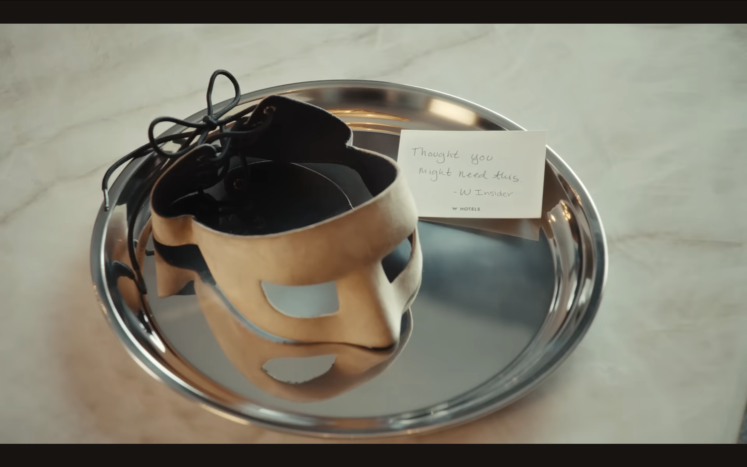 A metallic tray holds a pair of black and beige slippers shaped like a mask, with a note saying 'Thought you might need this - W Insider' next to it.