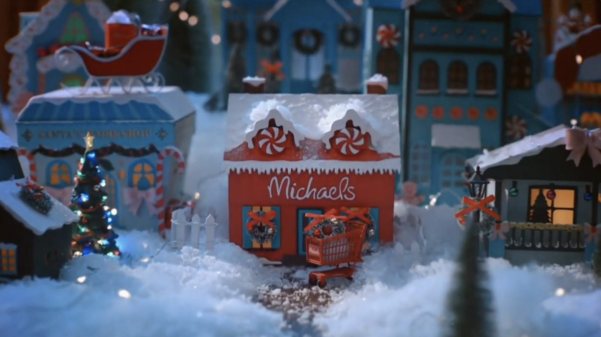 Miniature Christmas village scene with snow, decorated houses, a Christmas tree, and a Michaels store with a red storefront and peppermint swirl decorations.