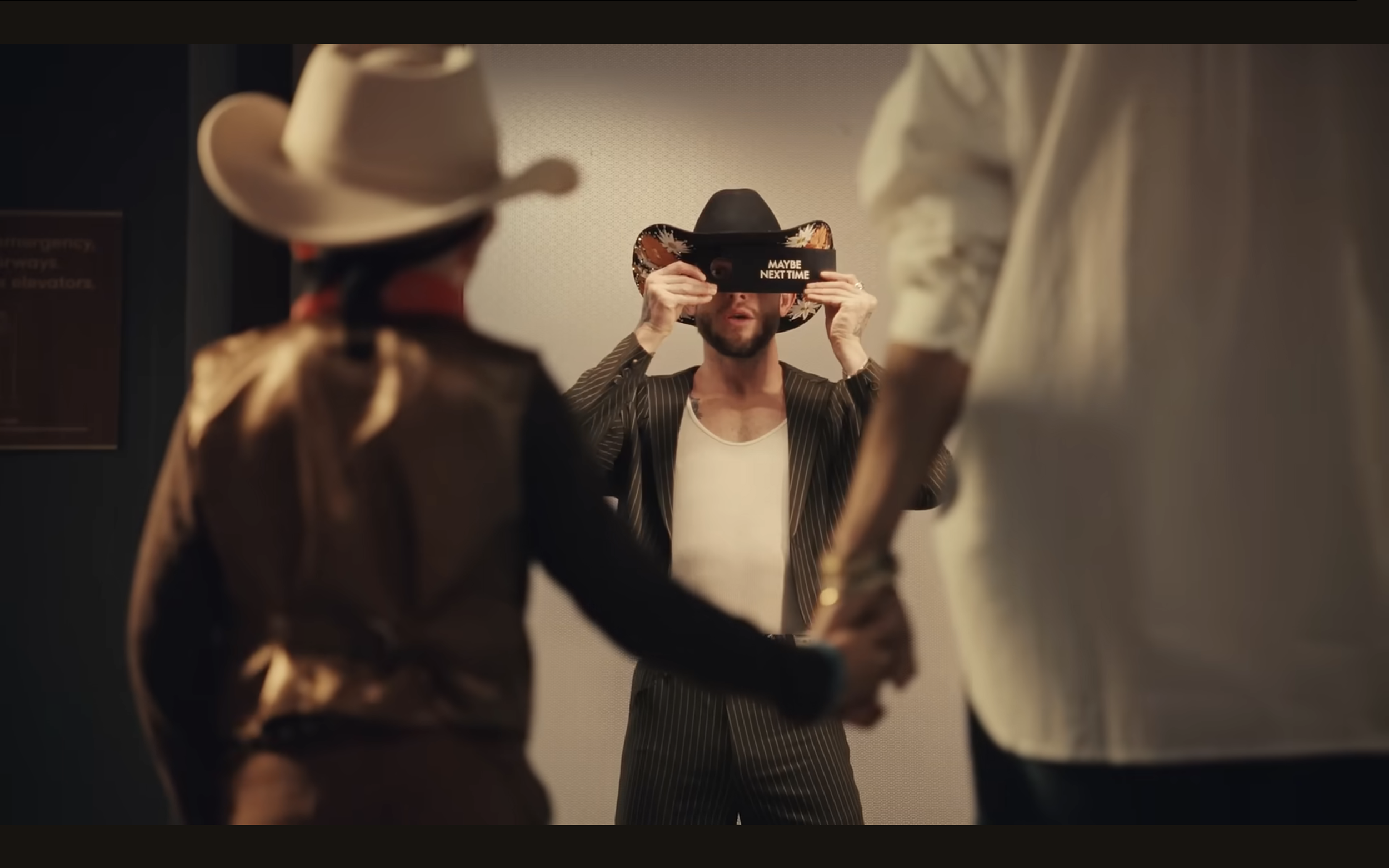 Man in pinstripe suit and cowboy hat holding a sign that says 'MAYBE NEXT TIME,' standing behind two people, with one of them in a cowboy hat.