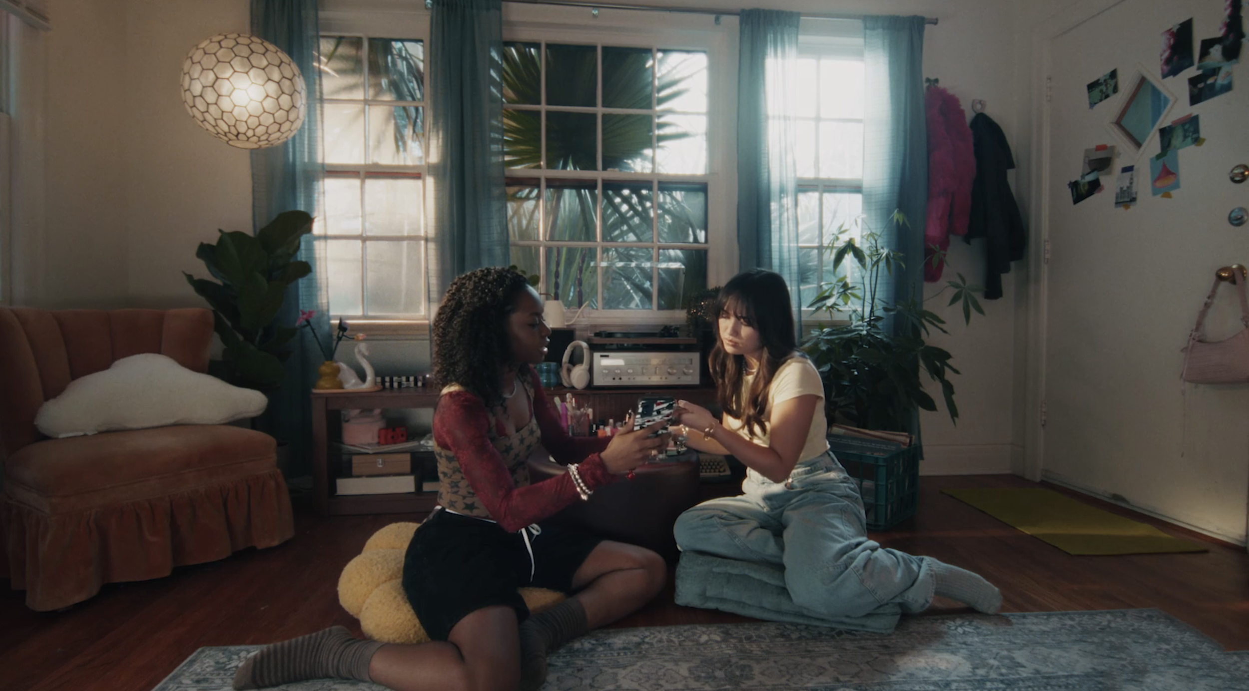 Two young women sitting on the floor in a cozy, well-lit living room, looking at a phone together.