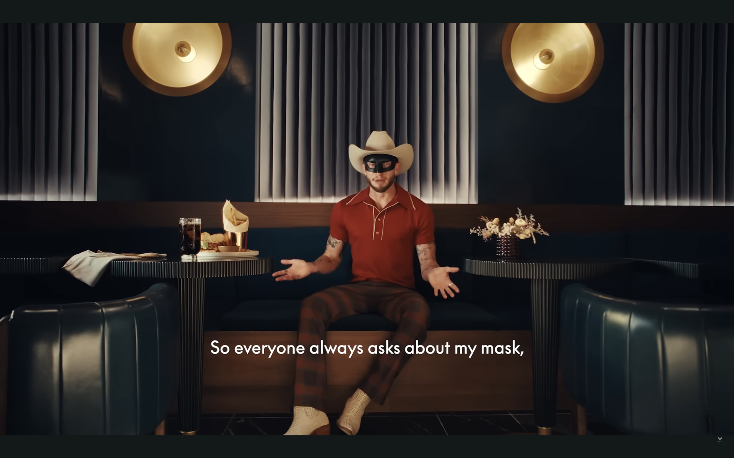 Man sitting in a restaurant booth wearing a cowboy hat and a mask, with a drink and food on the table and a bouquet of flowers on the other table, with a caption 'So everyone always asks about my mask'.