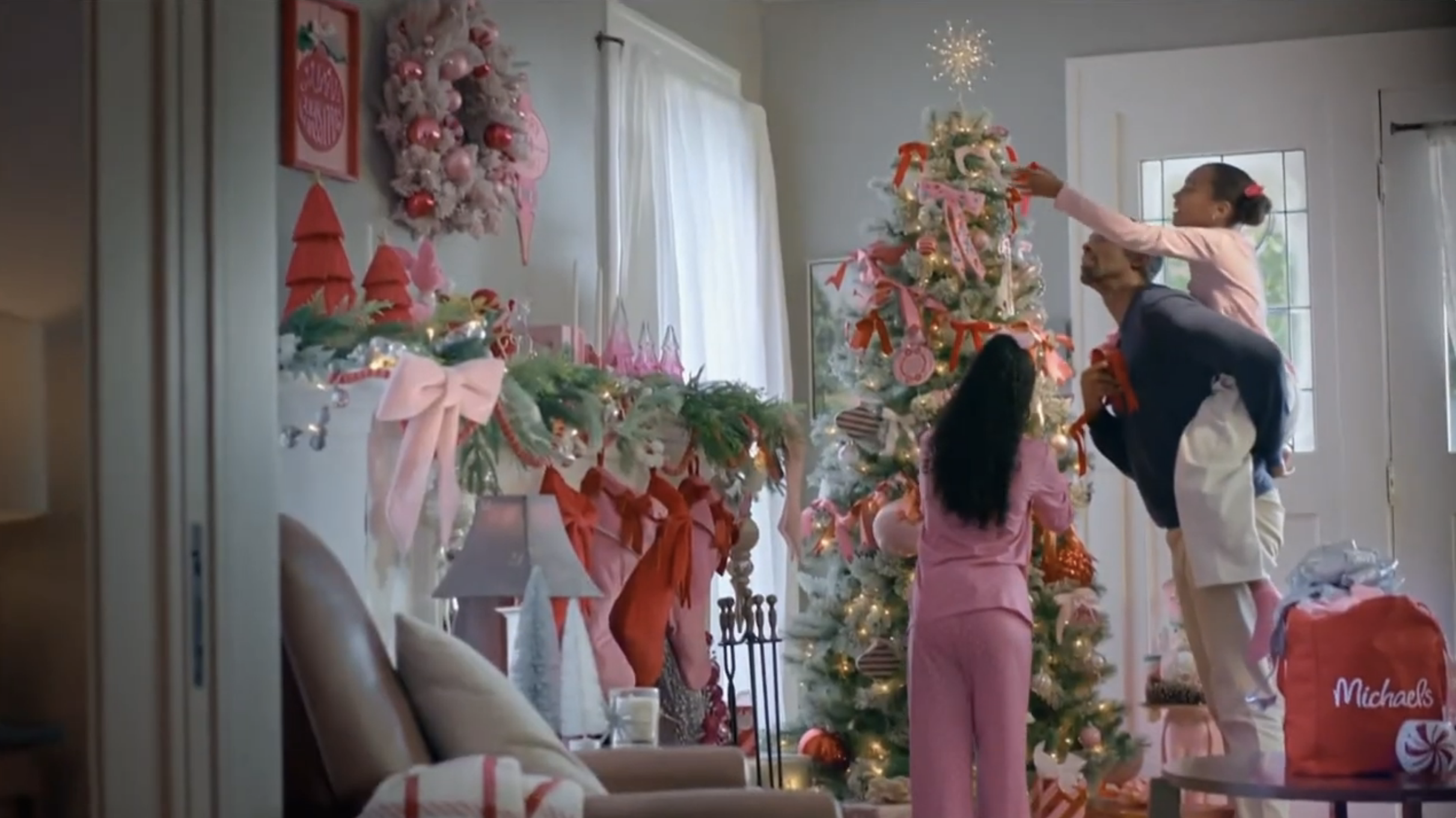 A family decorating a Christmas tree in a cozy, brightly lit living room with pink and red holiday ornaments, ribbons, and lights.