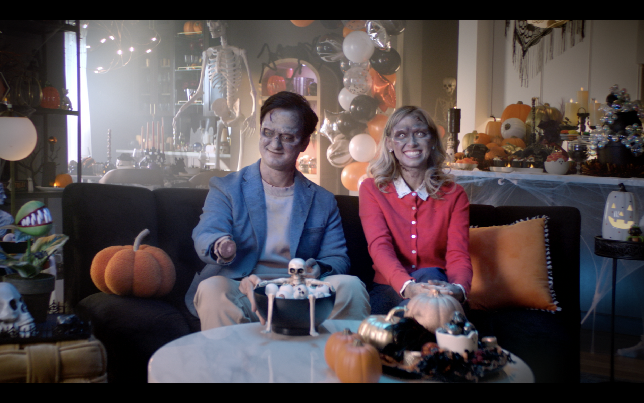 A man and woman dressed as zombies sit on a decorated Halloween couch. The room is filled with Halloween decorations including pumpkins, skulls, skeletons, and balloons.