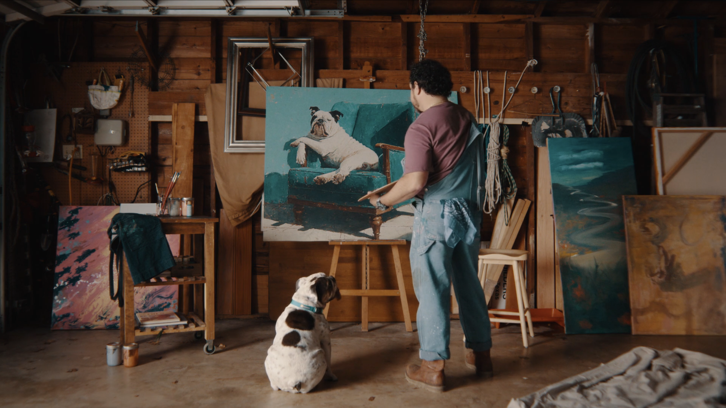 An artist painting a dog portrait in a wood-paneled studio. A dog sits on the floor watching. The studio has several paintings, art supplies, and tools.