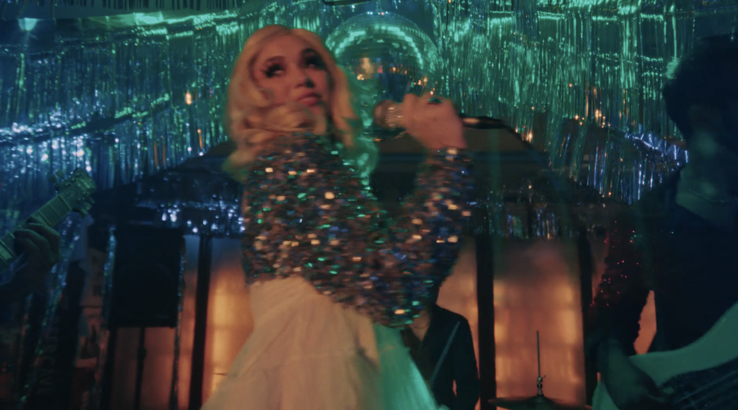 A woman singing into a microphone on stage, wearing a sparkly dress, with a reflective disco ball overhead and shiny silver streamers hanging in the background, while a man plays guitar beside her.