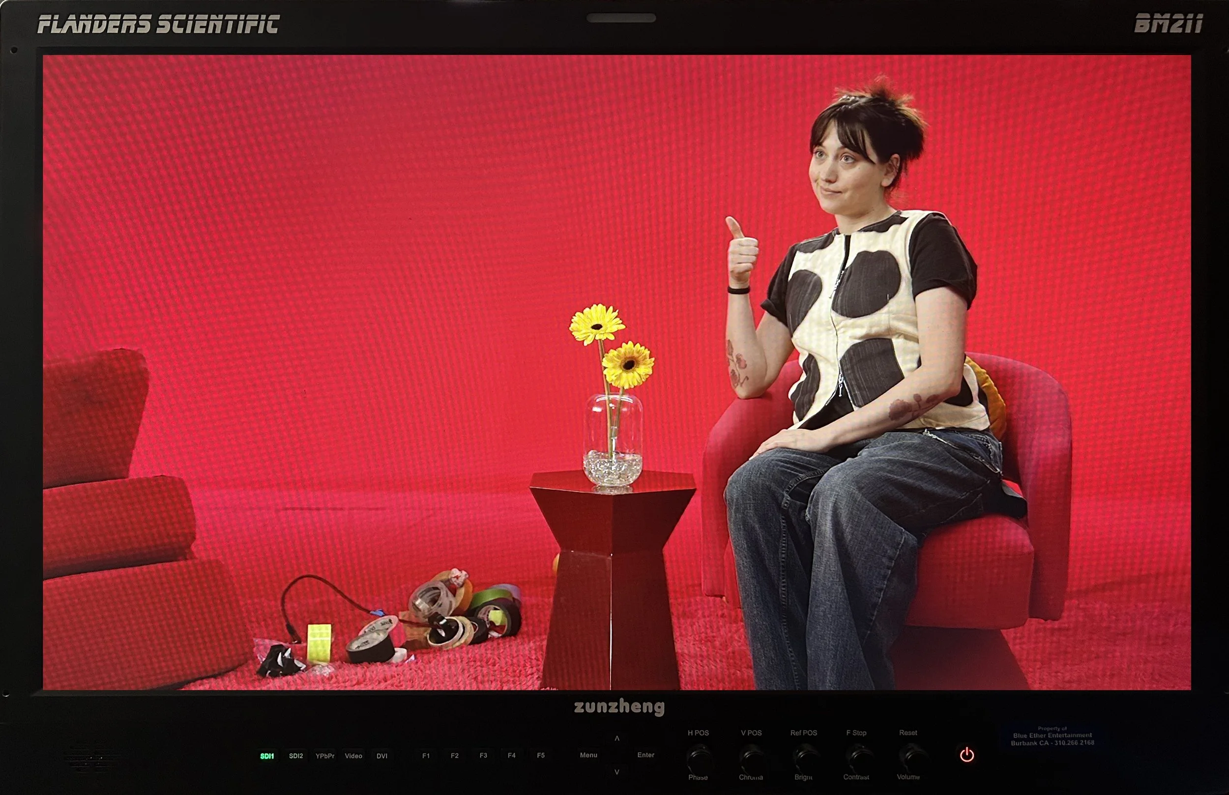 A woman sitting on a red couch with a red background, giving a thumbs-up gesture. A small table with yellow flowers in a glass vase is in front of her. There are various rolls of tape and tape dispensers on the floor to the left of the table.