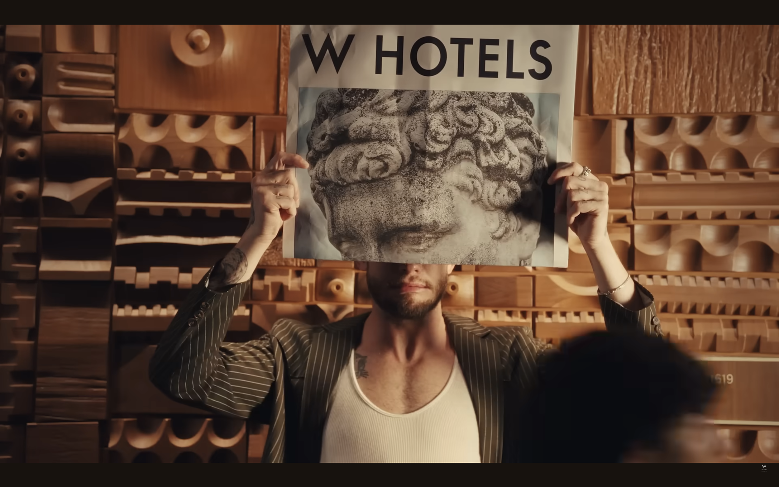 Man holding a poster of a stone sculpture's face above his face inside a room with wooden acoustic panels.
