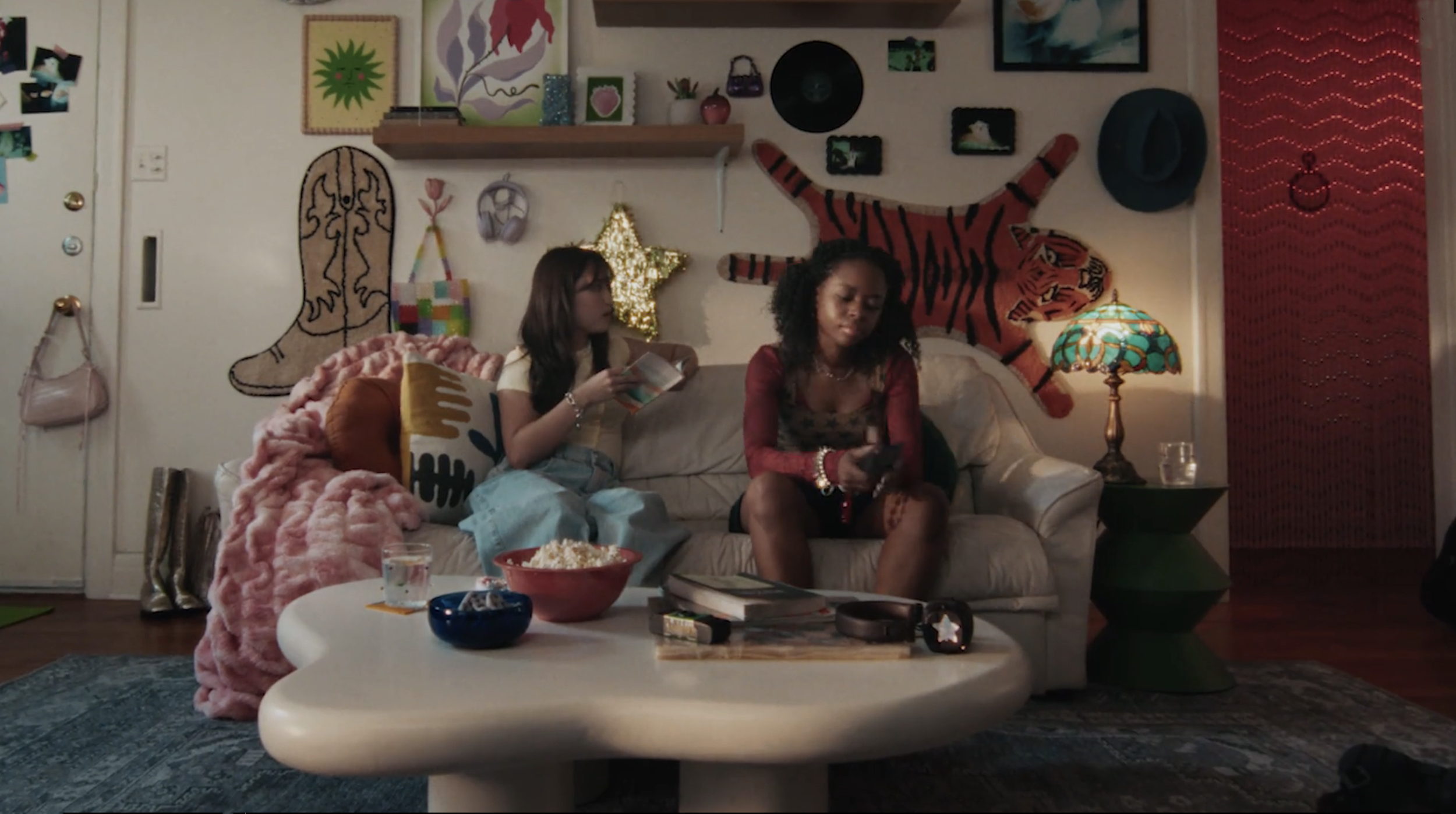 Two women sitting on a beige couch in a colorful, eclectic living room, one reading a book and the other looking at her phone, surrounded by decorative wall art, a lamp, and a coffee table with snacks and magazines.
