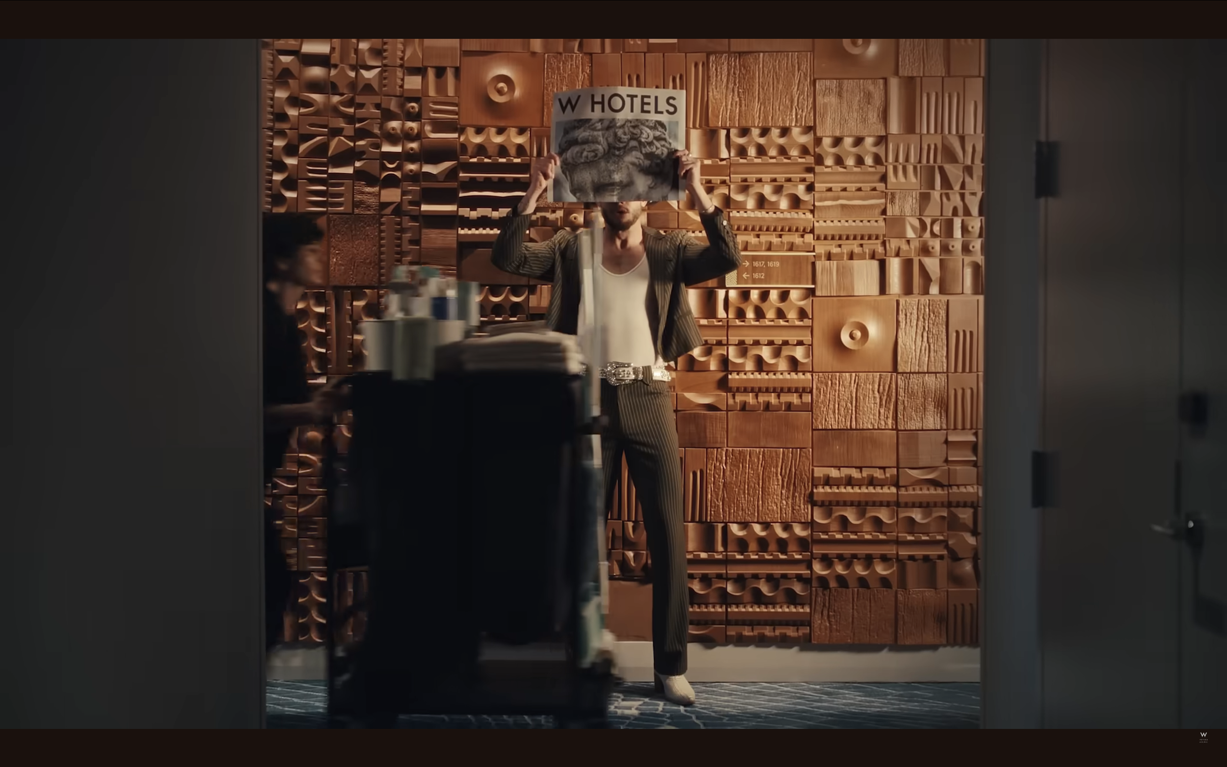 A man standing in front of a textured wooden wall, holding a magazine with the title 'W HOTELS' above his head. He is dressed in a pinstripe suit with a white shirt and wearing white shoes. There are some books and papers on a cart in the foreground,