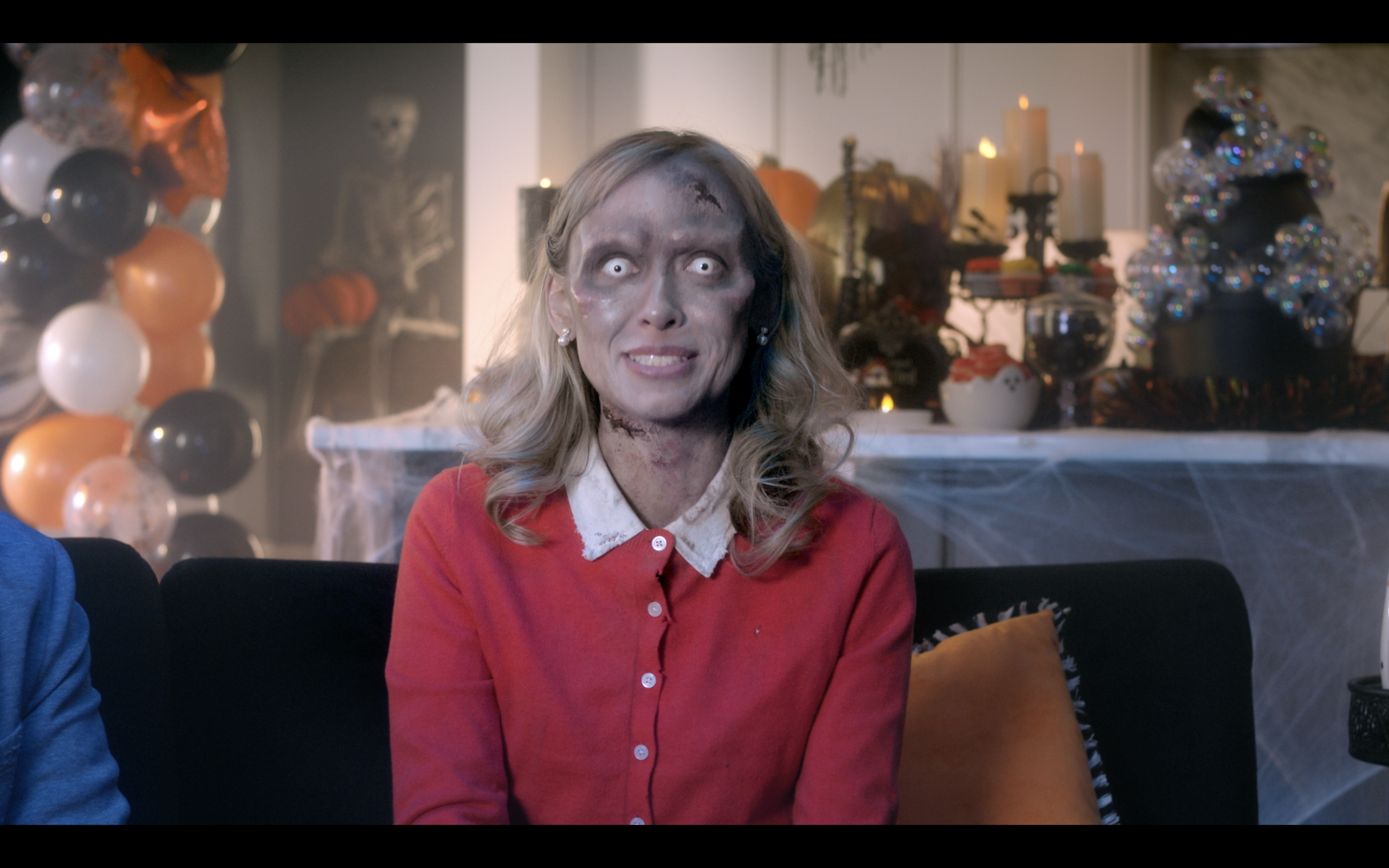 A woman with zombie makeup sitting on a couch during Halloween, with a spooky decorated background including balloons, pumpkins, skeletons, and cobwebs.