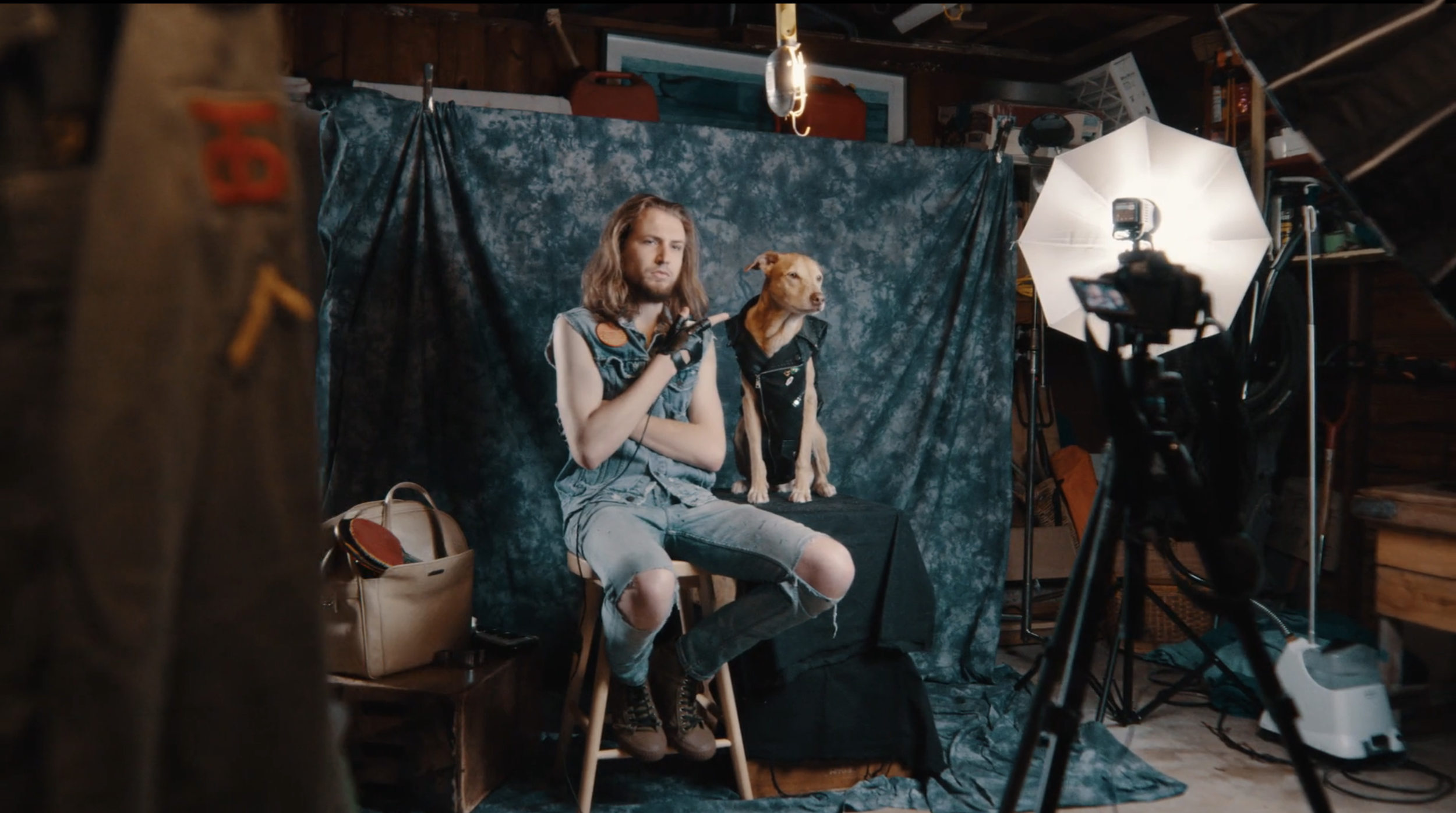 A young man with long hair sitting on a stool on a photography set, holding a small tool in his right hand, with a dog sitting beside him. The set has a dark patterned backdrop, studio lighting equipment, and various photography props around.