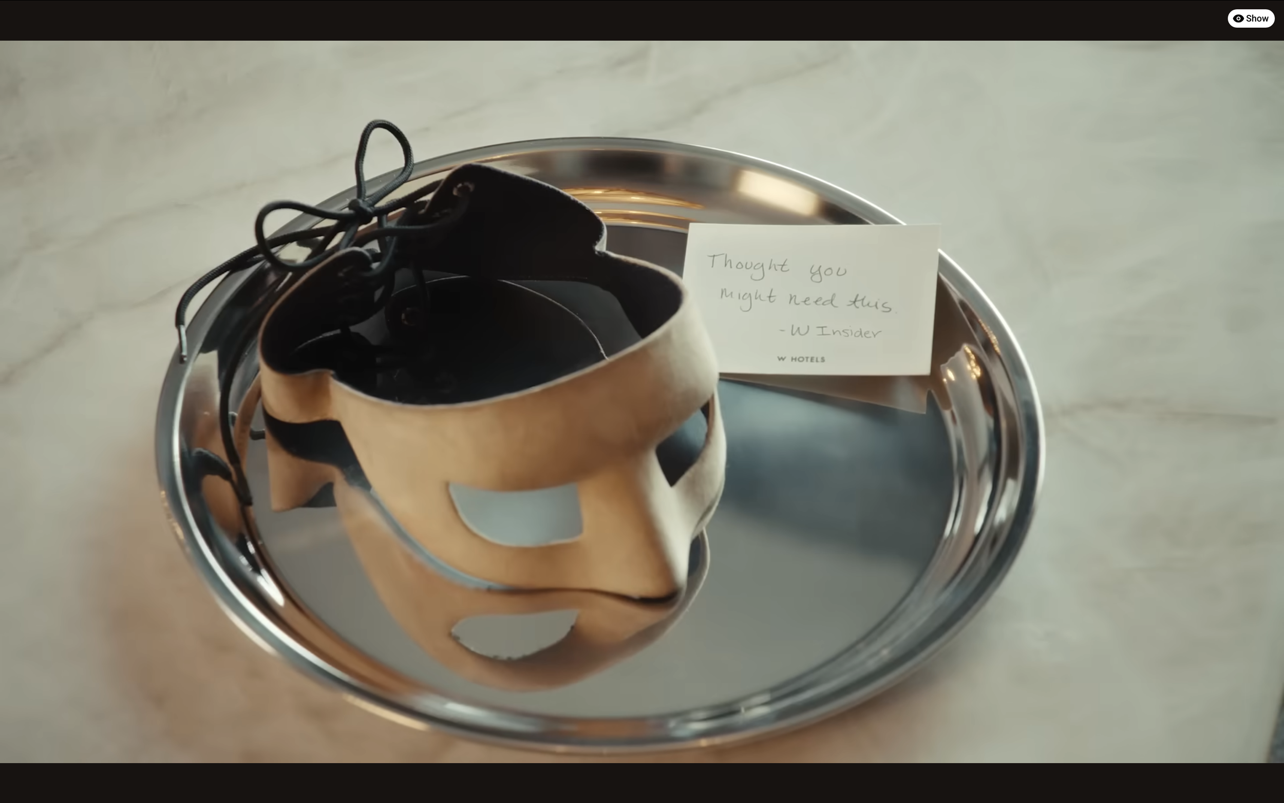 A silver tray with a black and gold masquerade mask, a pair of black shoes, and a small note that reads, "Thought you might need this. - W Insider" on a marble surface.