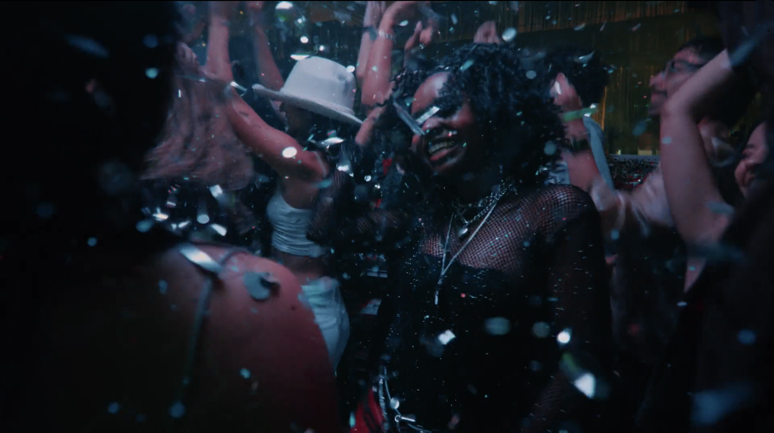 People dancing at a party with confetti falling, showing a woman with curly black hair and a fishnet top smiling.