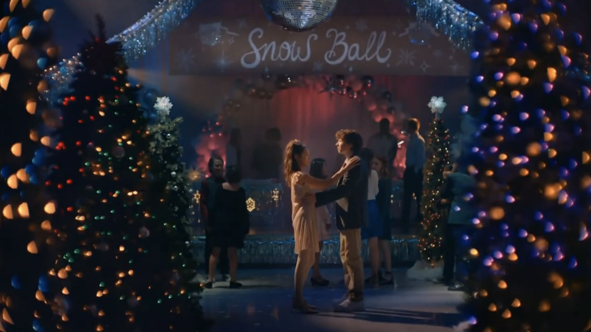 Couple dancing at a Christmas-themed event called Snow Ball, decorated with Christmas trees and colorful lights.