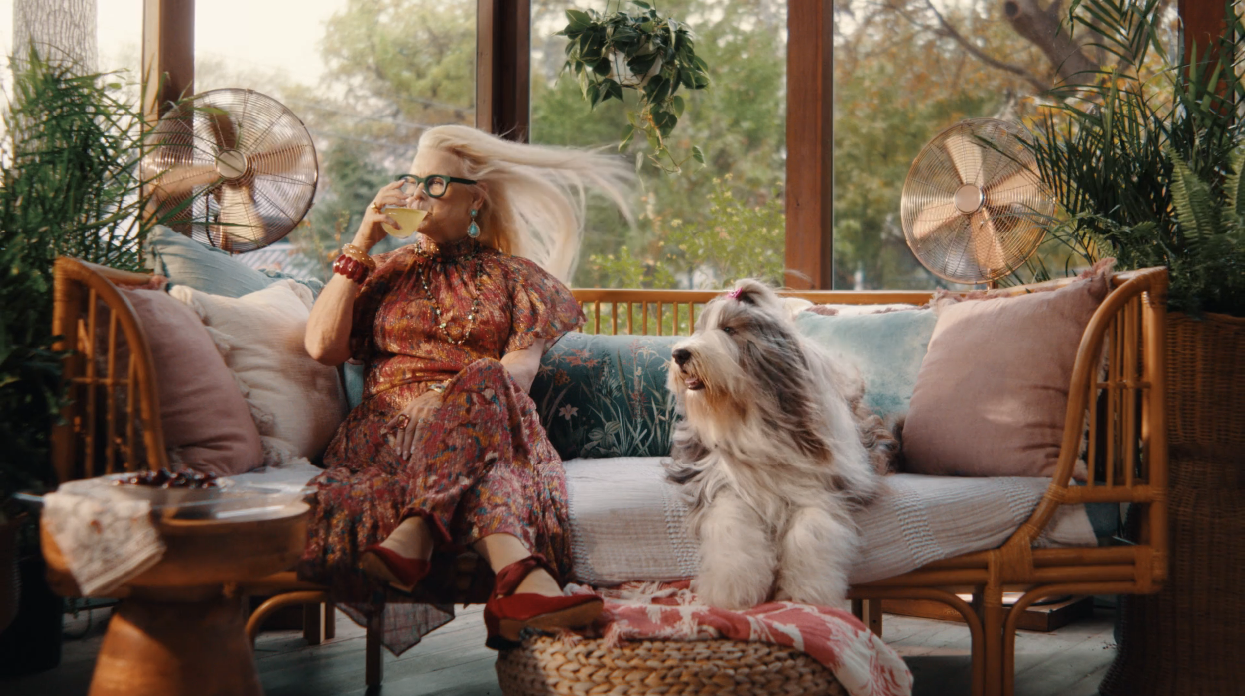 An elderly woman with long blonde hair, glasses, and colorful jewelry, wearing a floral dress, is sitting on a wicker couch drinking from a cup. She has her hair flowing in the air and is surrounded by green plants and vintage fans. She is accompanie