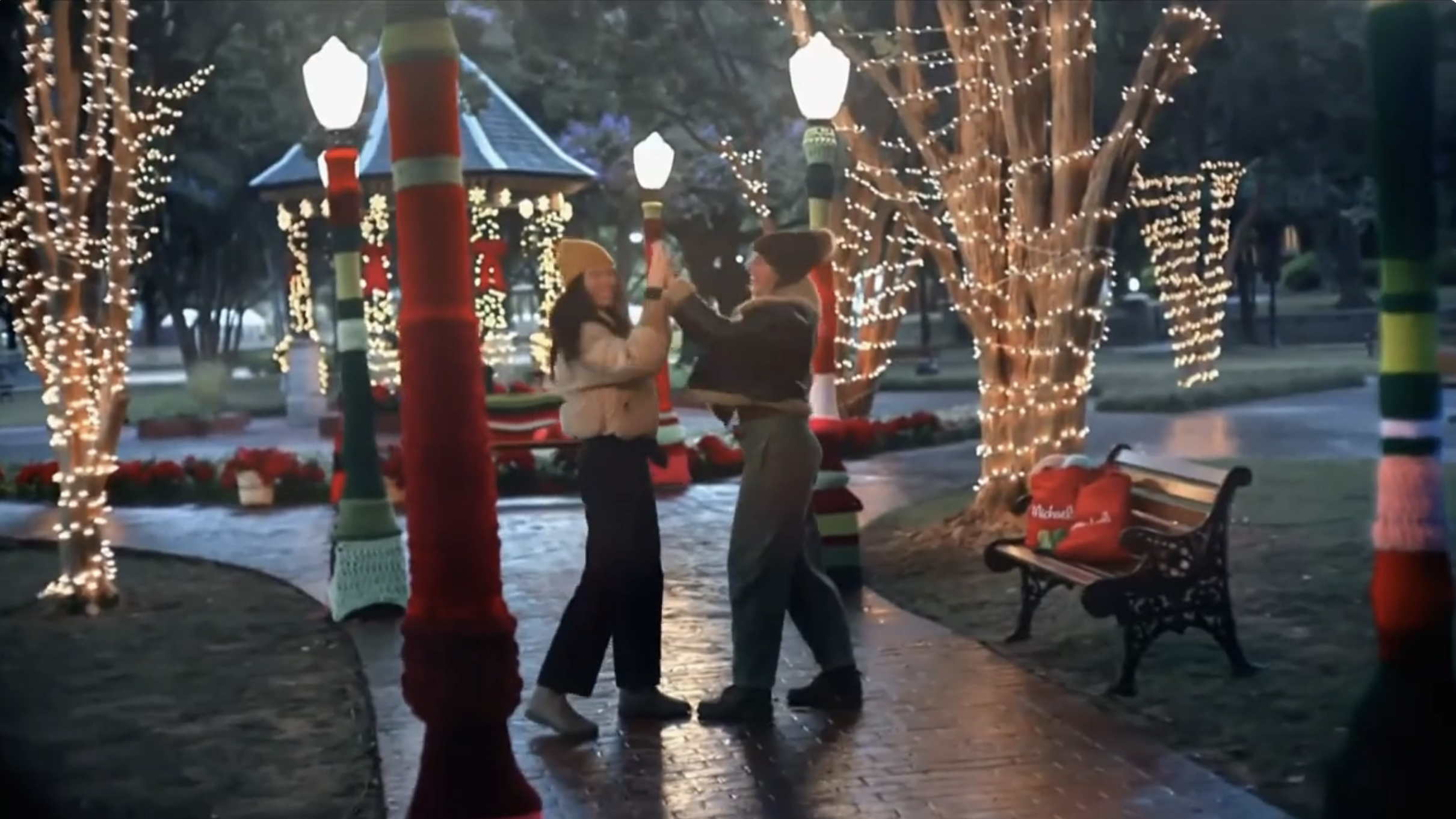 A couple dancing together in a park decorated with Christmas lights and ornaments, with trees wrapped in fairy lights and a gazebo in the background.