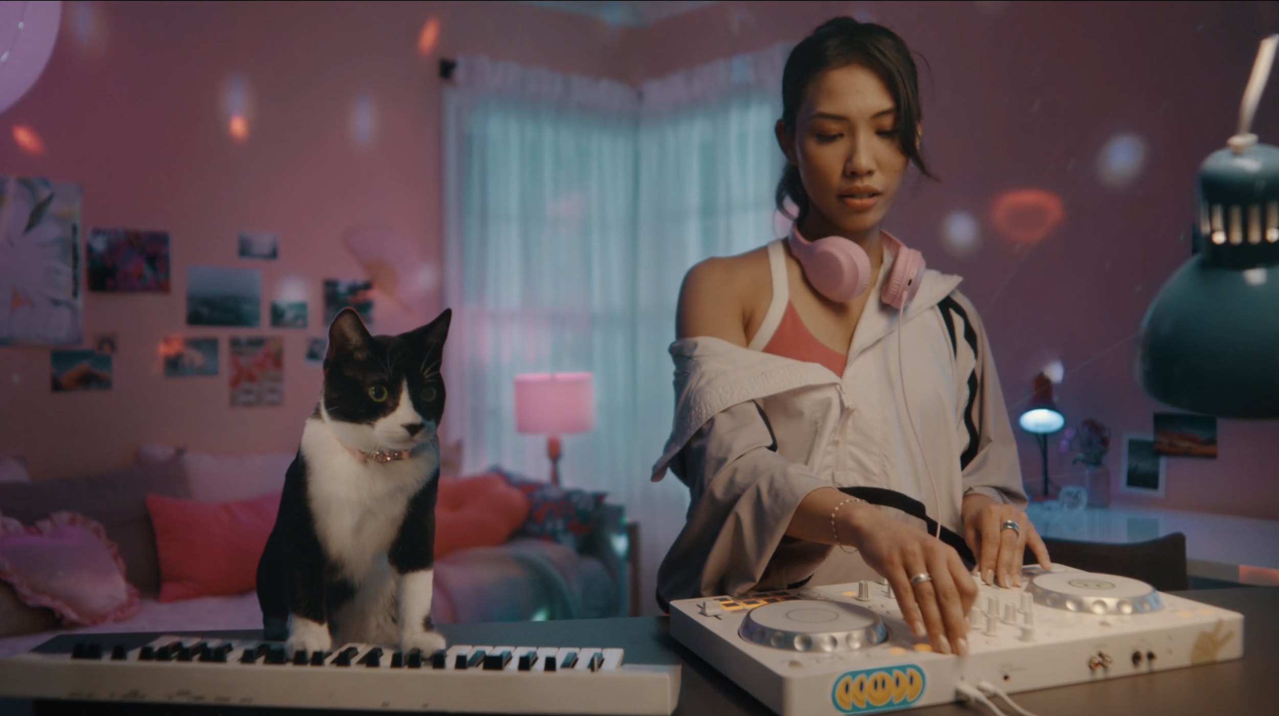 A woman with short dark hair wearing a pink tank top, white hoodie, and pink headphones, DJing on a white turntable in a colorful, cozy room with pink walls, a black-and-white cat sitting on a keyboard, and various decorative items and photos on the 