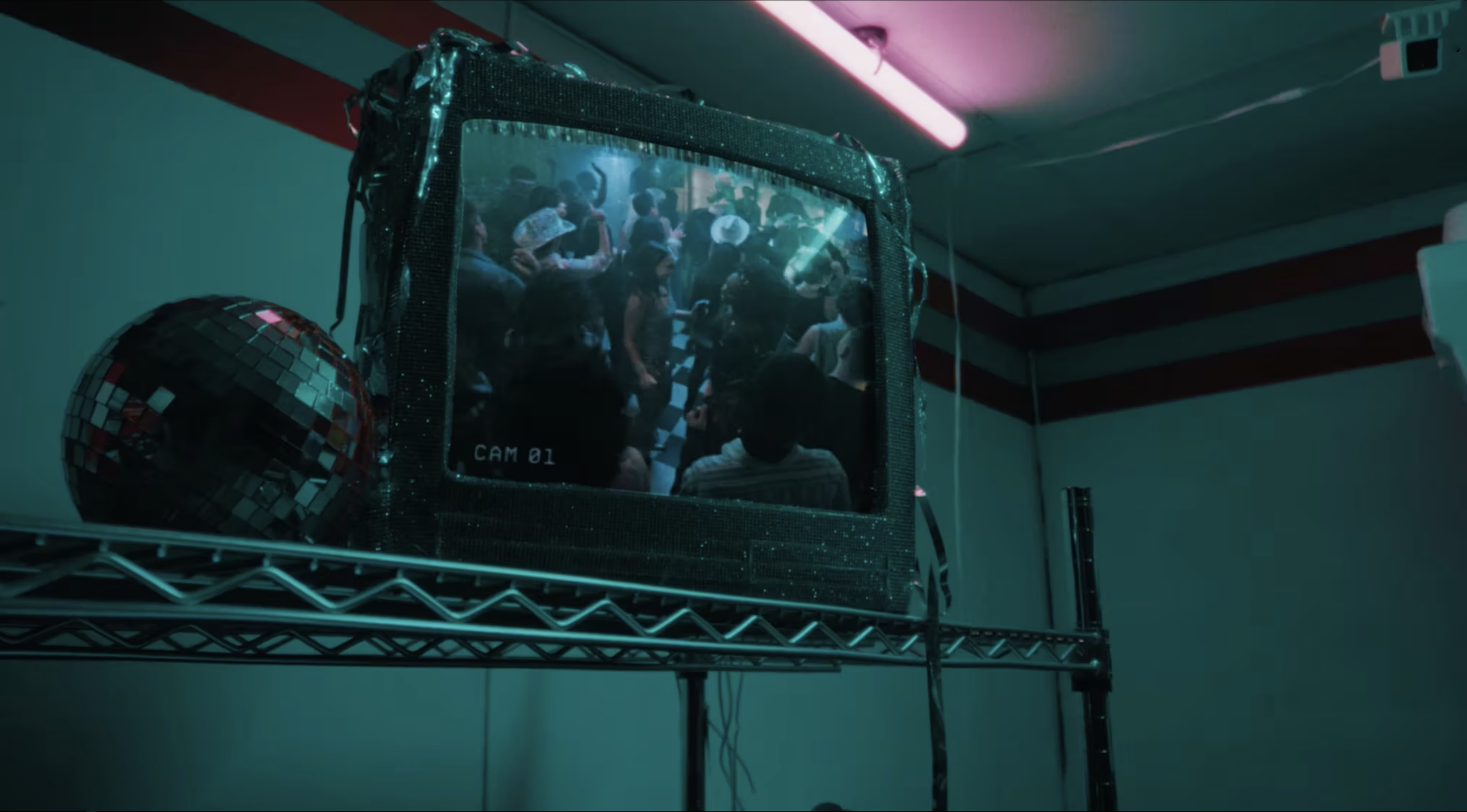 A crowded indoor party scene viewed through a TV or monitor screen on a metal shelf, with a disco ball nearby and dim, colorful lighting.