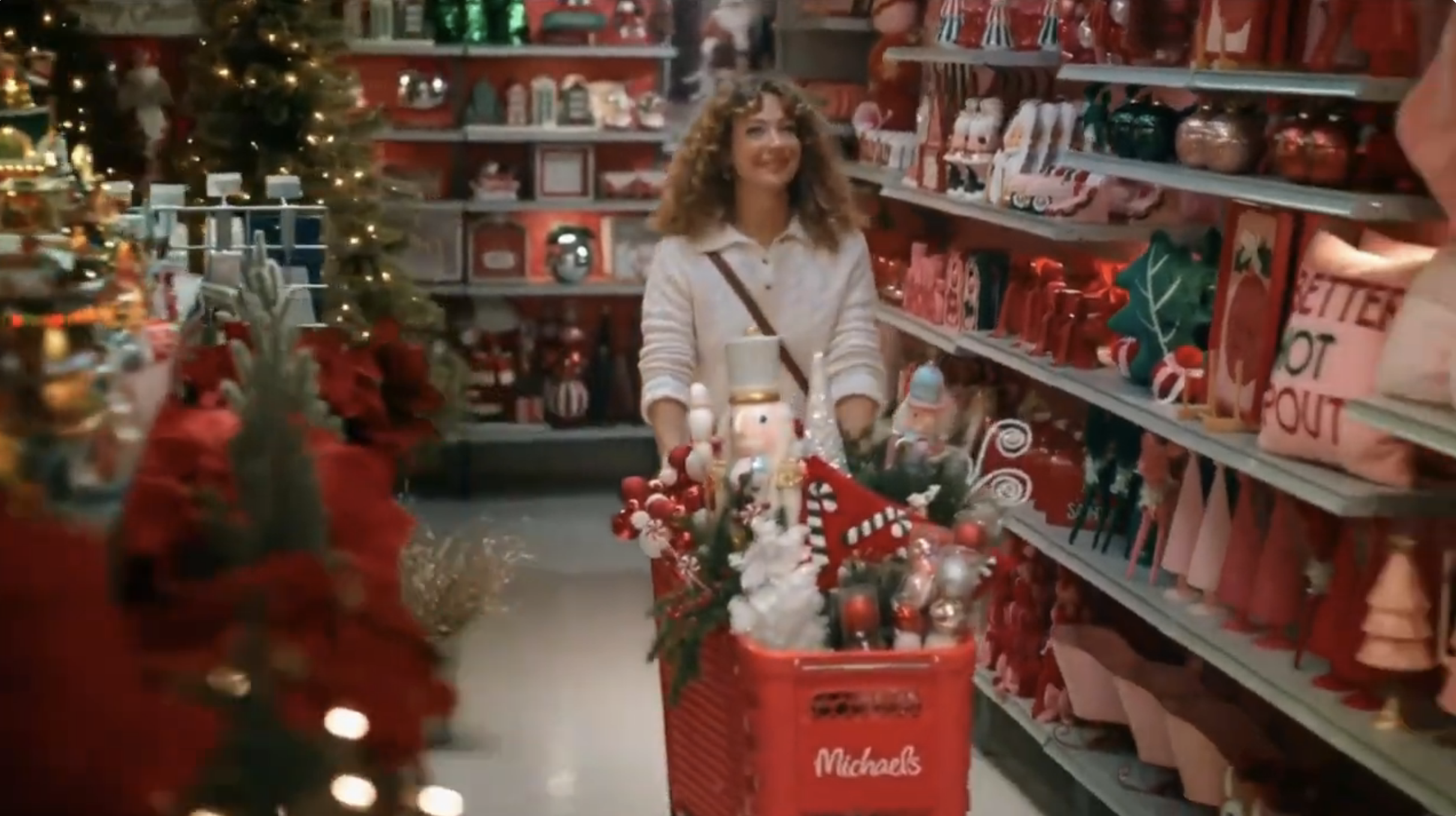 A woman shopping for Christmas ornaments at Michaels store, pushing a red shopping cart filled with holiday decorations, in front of shelves stocked with Christmas decorations and a decorated Christmas tree.