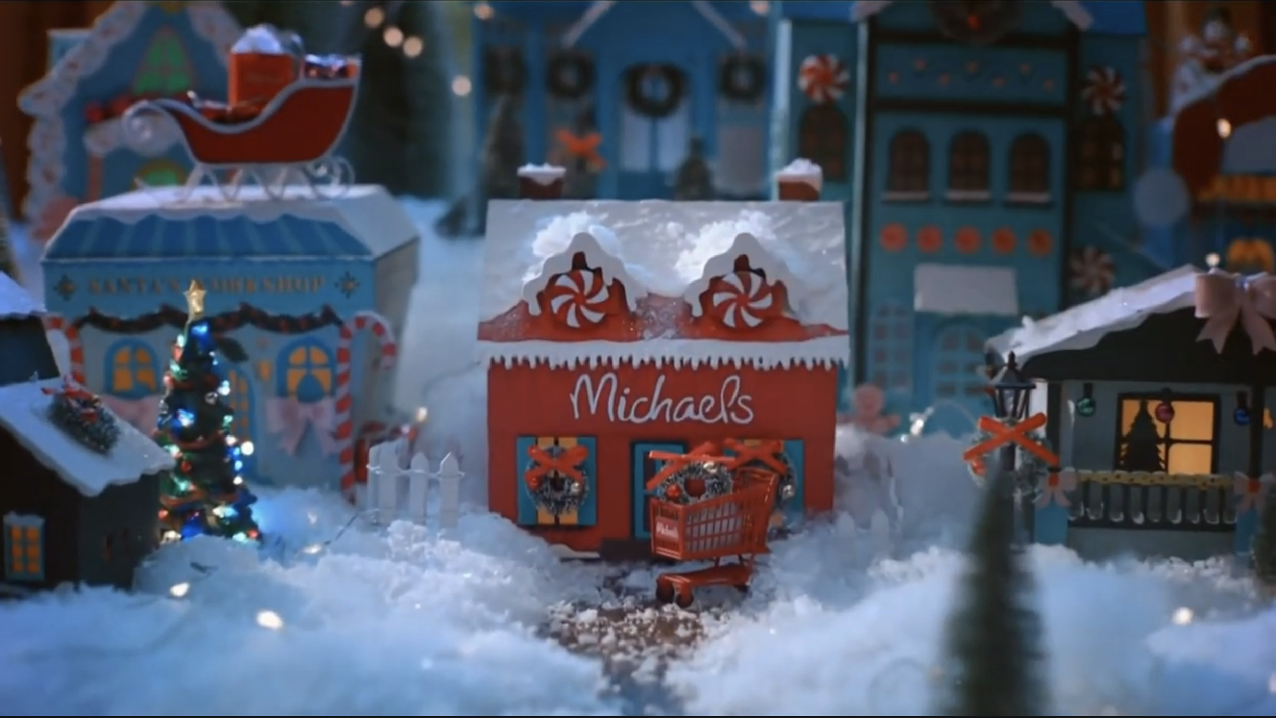 Miniature Christmas scene with a red Michaels store, small decorated Christmas tree, toy shopping cart, and various decorated buildings, all covered in snow.