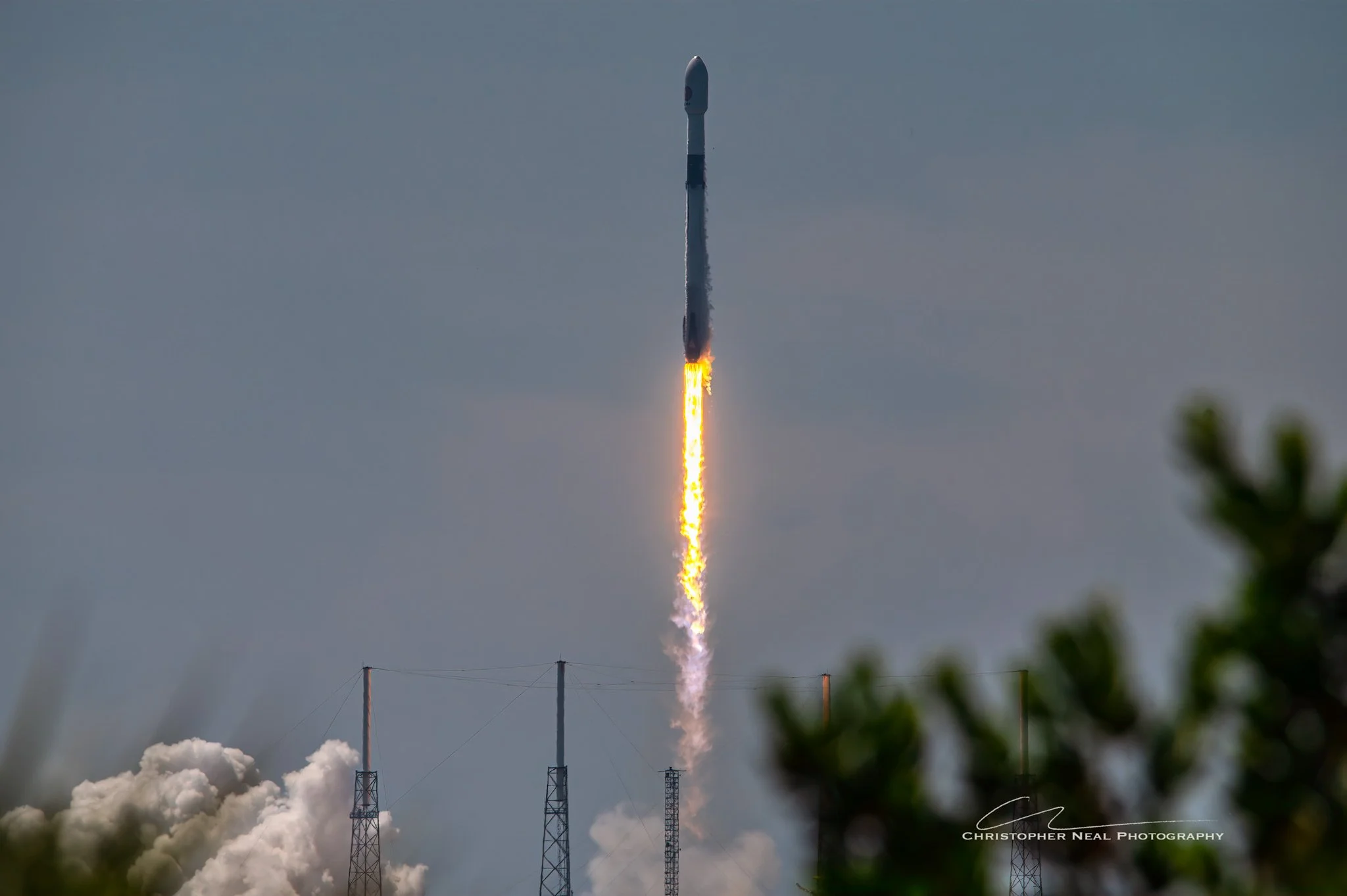 A rocket launching into the sky with flames and smoke under a cloudy sky, with structures and power lines in the foreground.