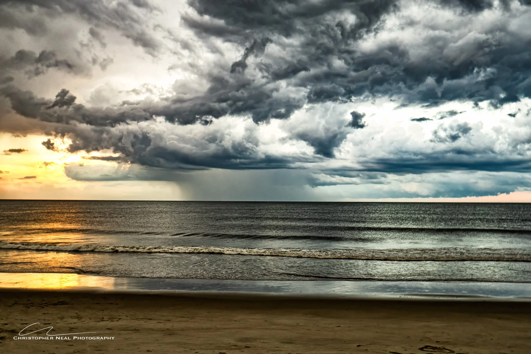 Cloudy sky over the ocean at sunset with dark, heavy clouds and choppy waves, beach in the foreground.