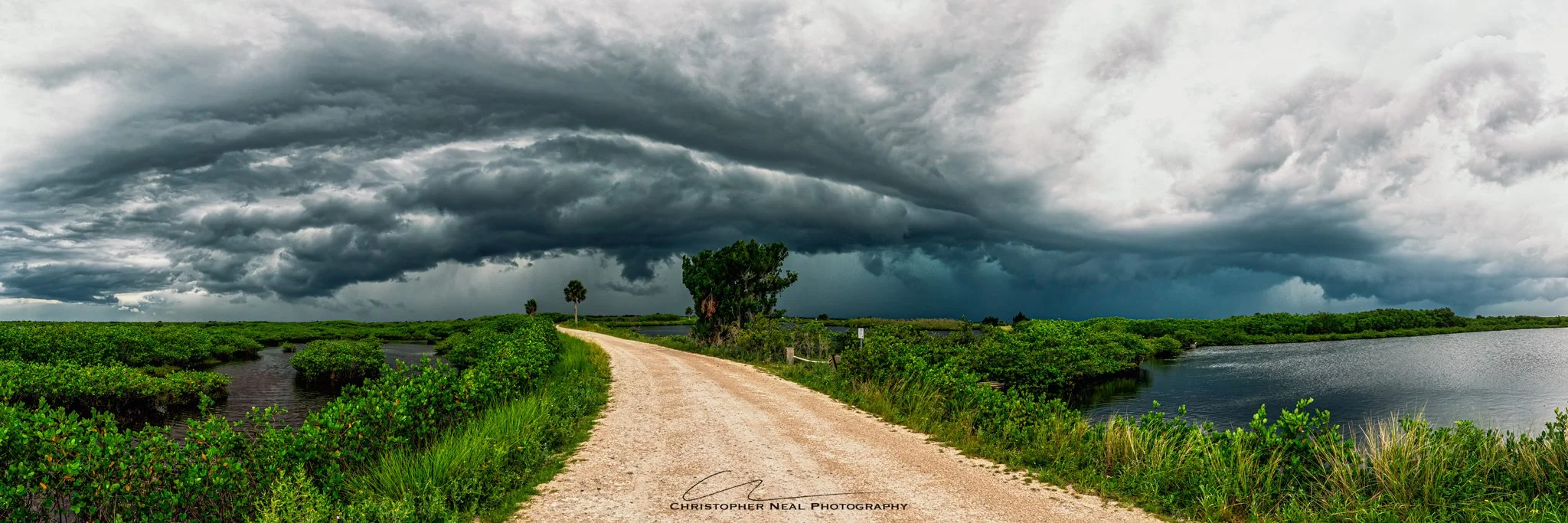 A dirt pathway runs through a lush green marshland with water bodies on both sides, under a dark, stormy cloudy sky.