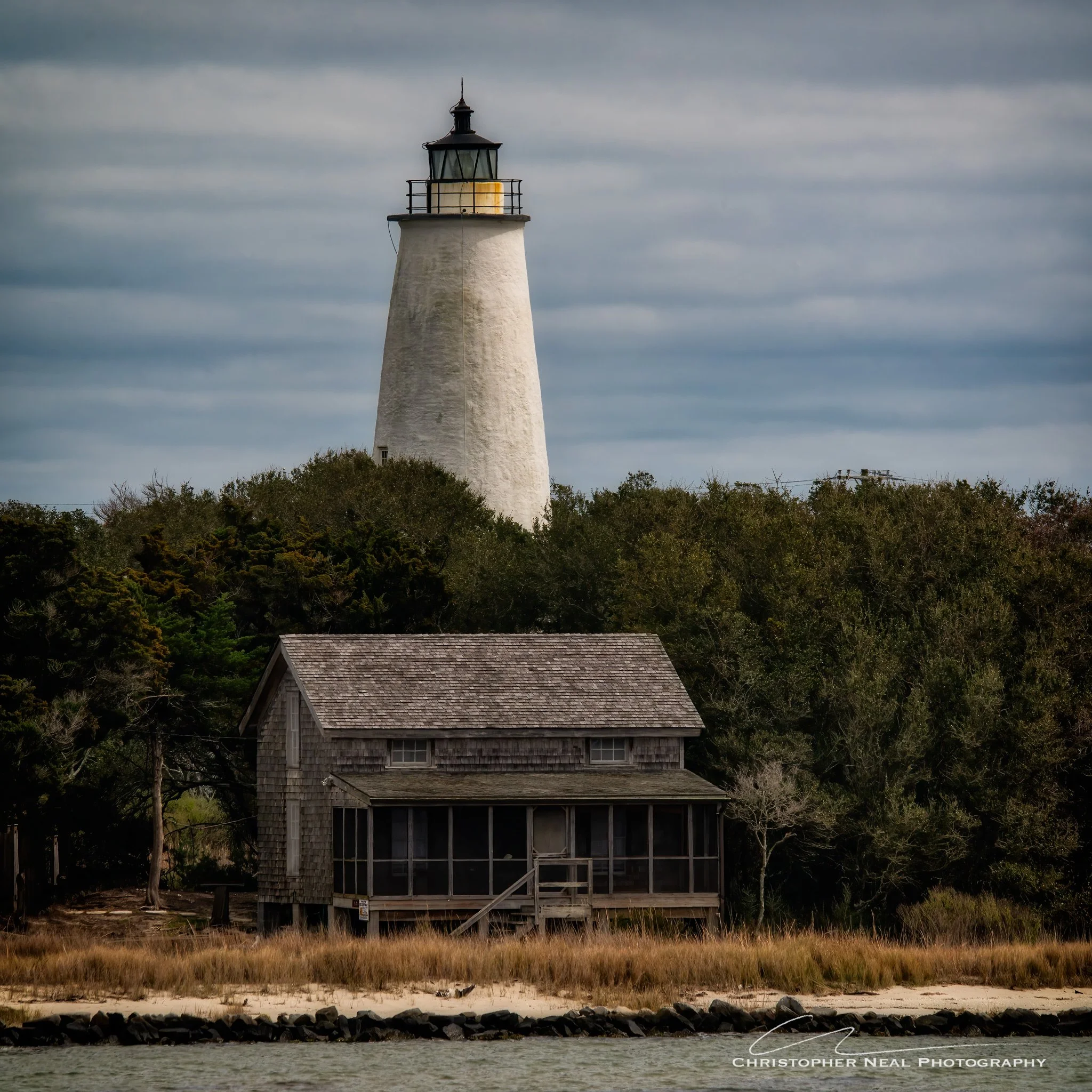 A lighthouse perched on a hill with trees, a wooden house with a shingled roof in the foreground, and a body of water at the bottom of the image. Overcast sky with gray clouds.