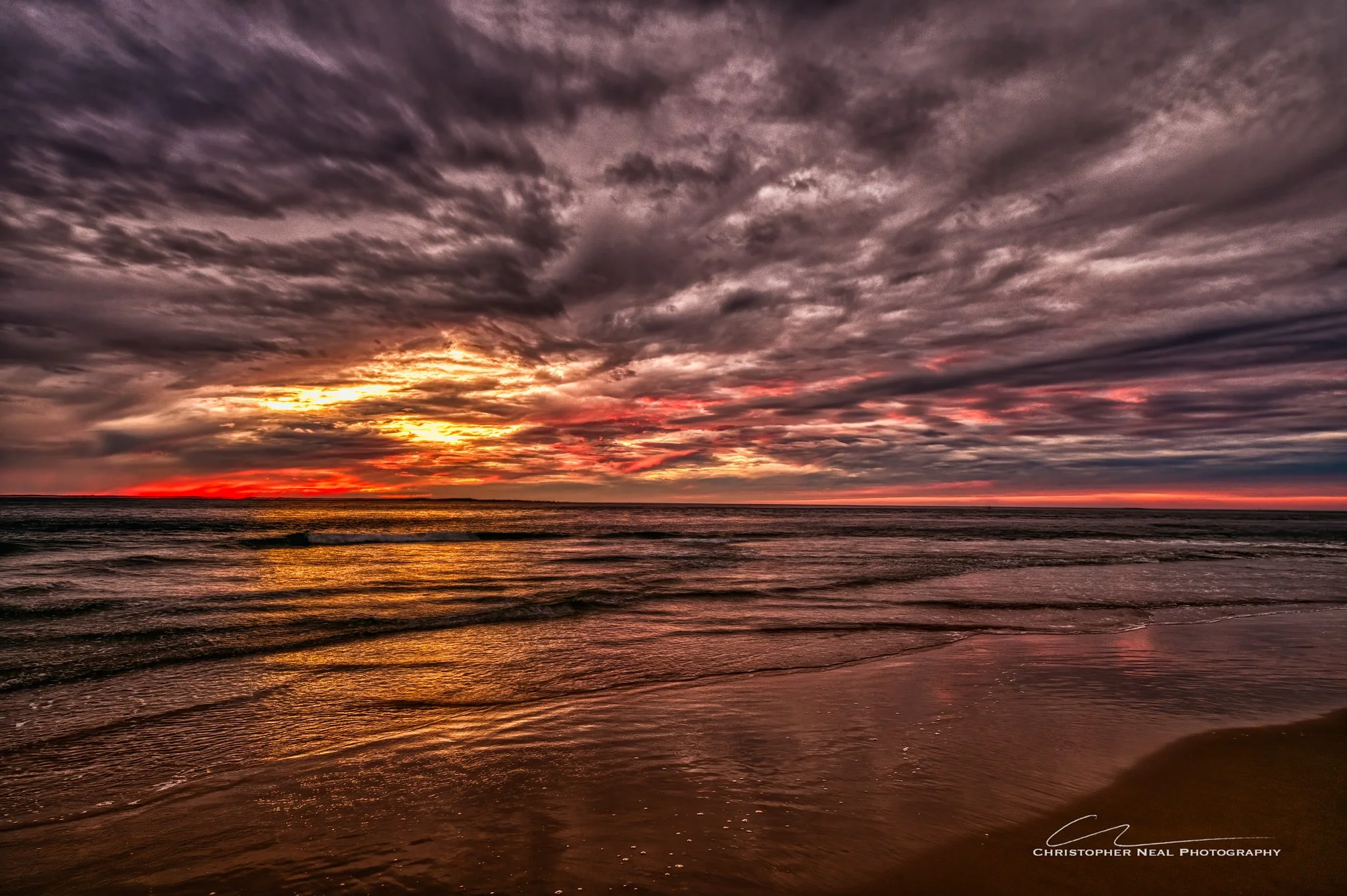 A sunset over the ocean with a cloudy sky, where the sun's rays pierce through the clouds, casting a golden and pink reflection on the water and wet sand at the beach.