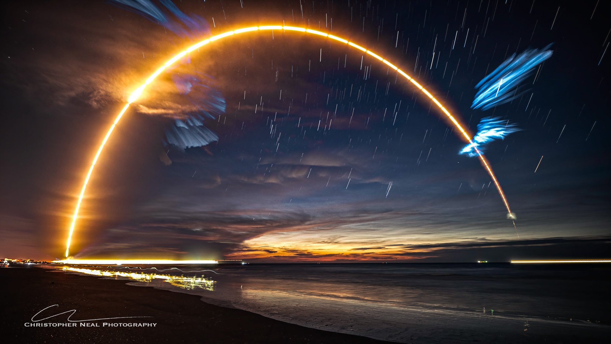 A time-lapse photograph of a rocket launch showing a bright, arched trail in the night sky, stars, clouds, and reflections on the water below.