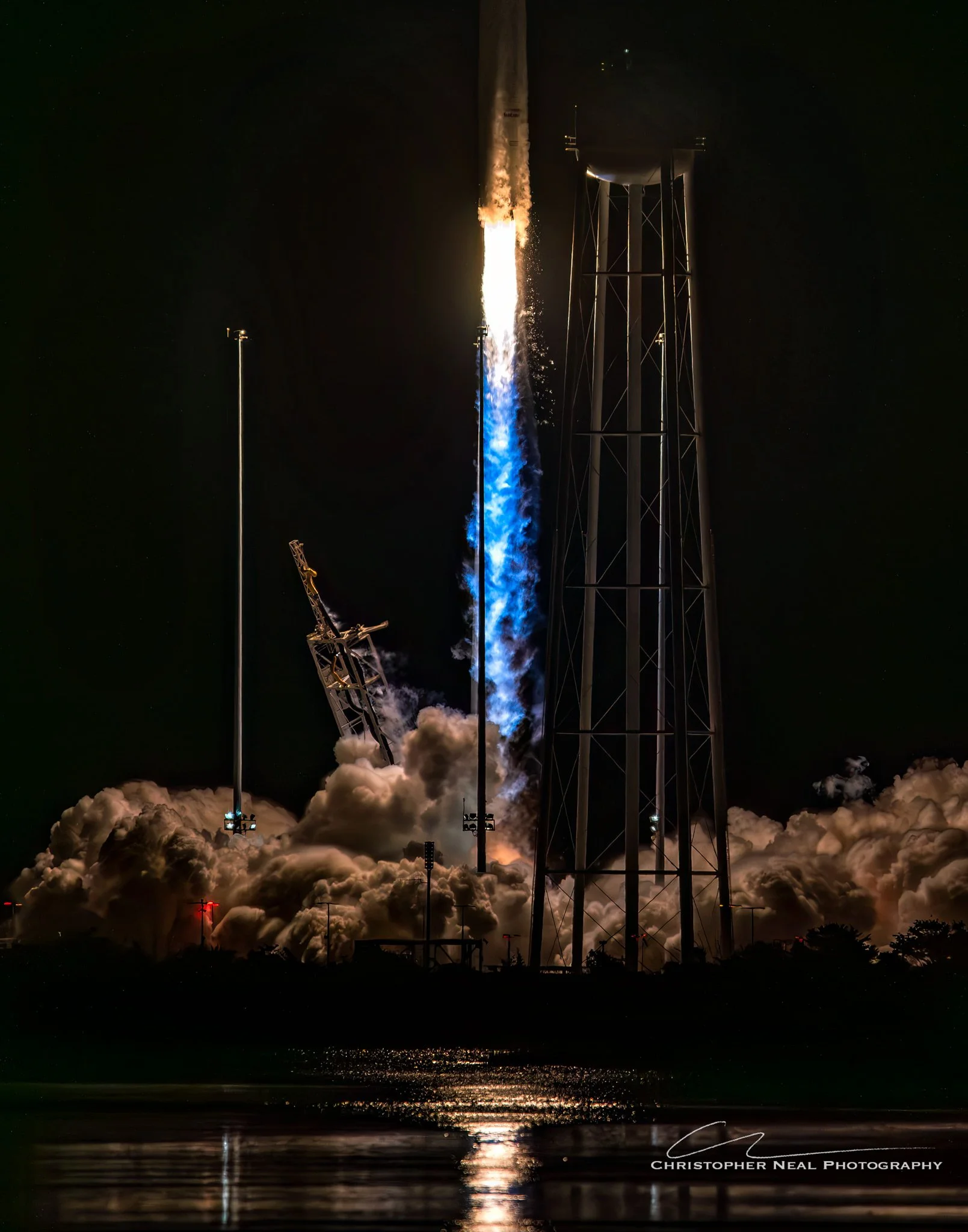 Nighttime rocket launch with flames and smoke, standing on a launch pad, reflected in water, with a dark sky and water below.