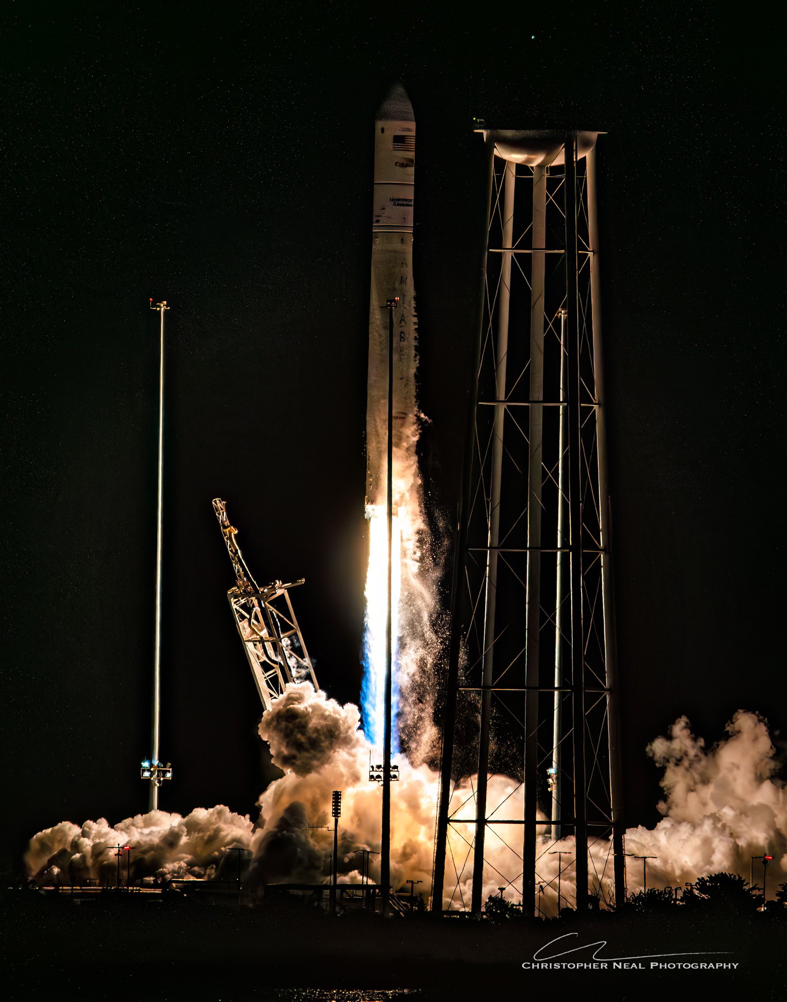 Photo of a rocket launch at night with a bright trail of flames and smoke, surrounded by a dark sky filled with stars, and some lit structures on the ground.