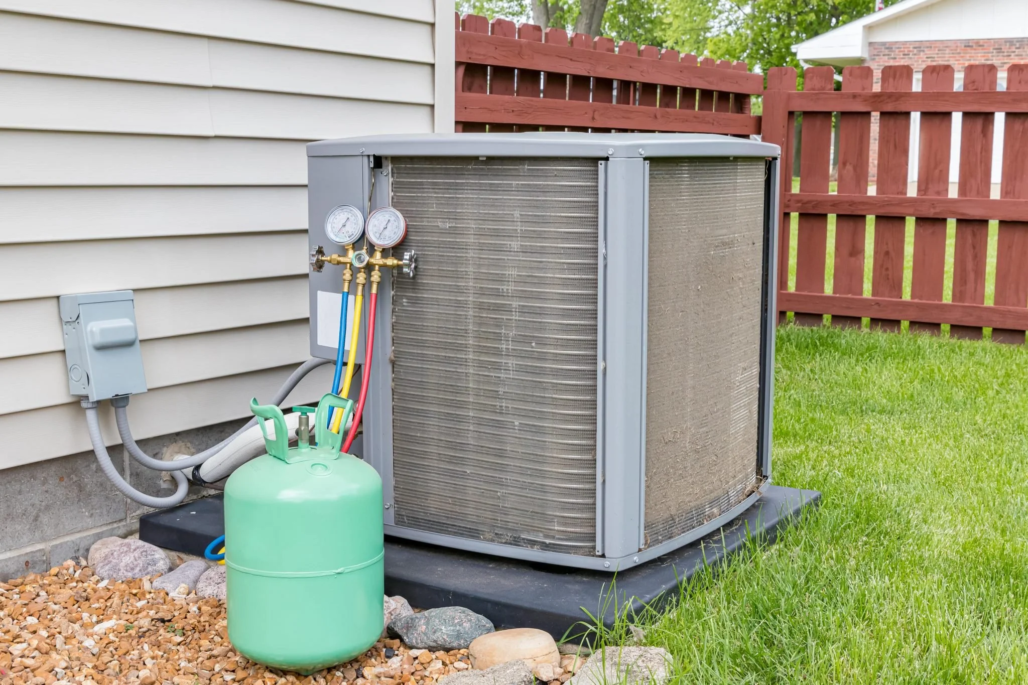 Central air conditioning unit outdoors next to a house with wiring and gauges, with a green refrigerant tank nearby, on a black pad surrounded by gravel and grass, with a red wooden fence and green trees in the background.