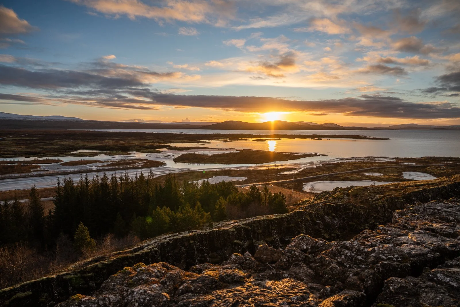 Sunset over a landscape with water, lakes, and trees, viewed from a rocky elevation.