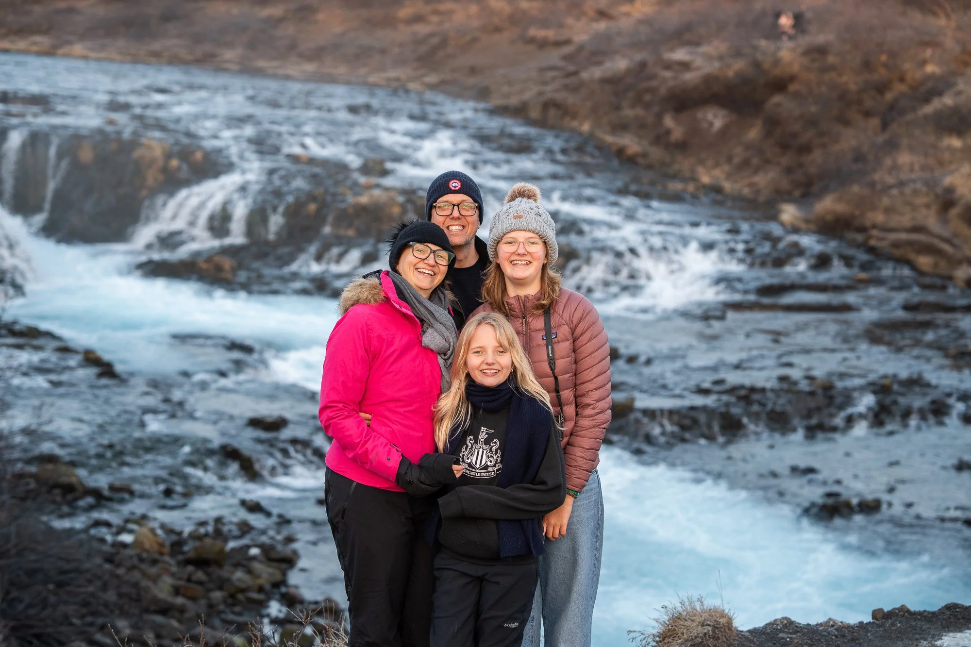 Family of five in outdoor winter clothing, standing near a rushing waterfall with rocky terrain in the background during daytime.